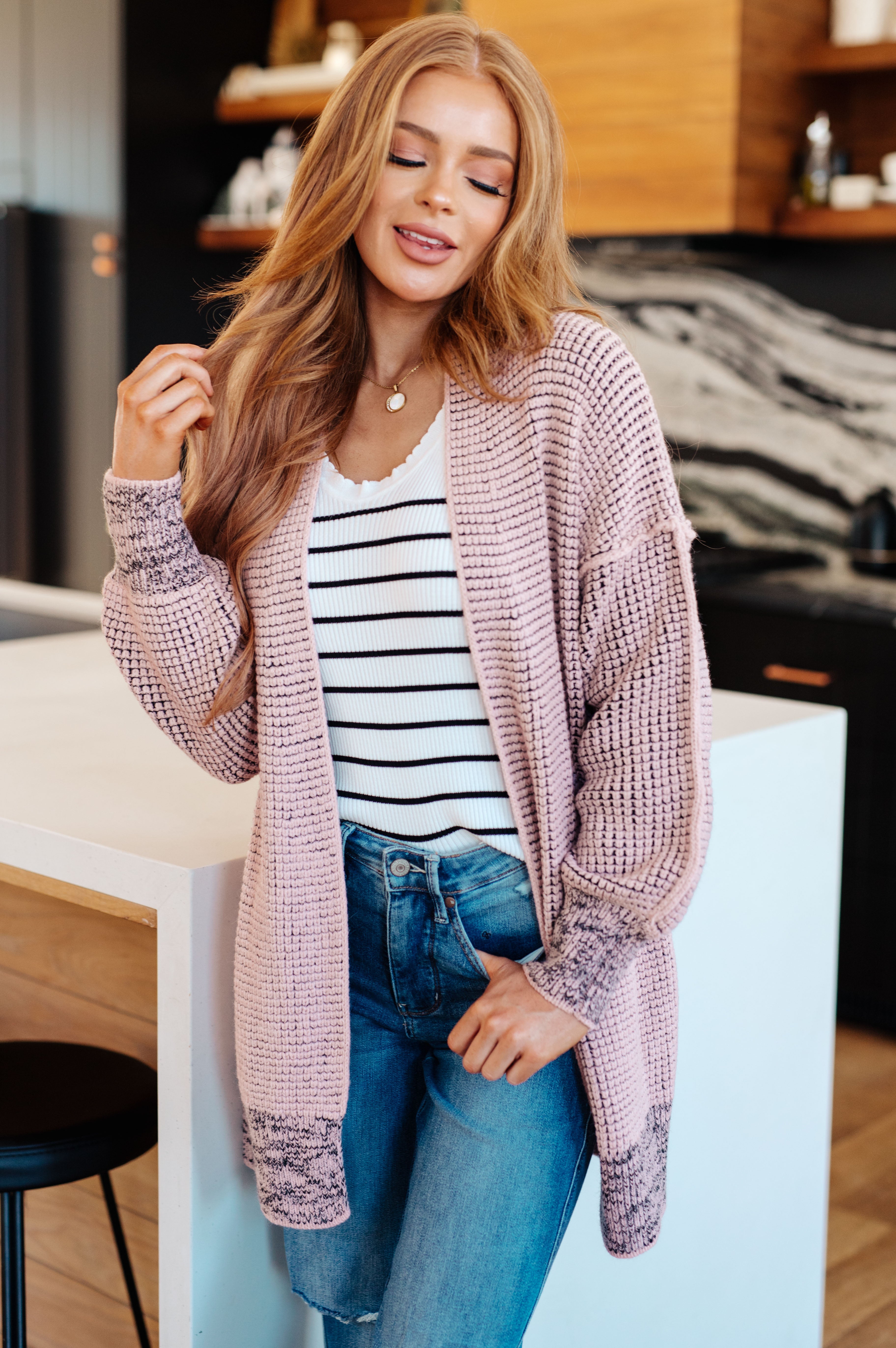 Woman wearing mauve knit cardigan over striped white top and jeans, standing in modern kitchen with warm wood accents