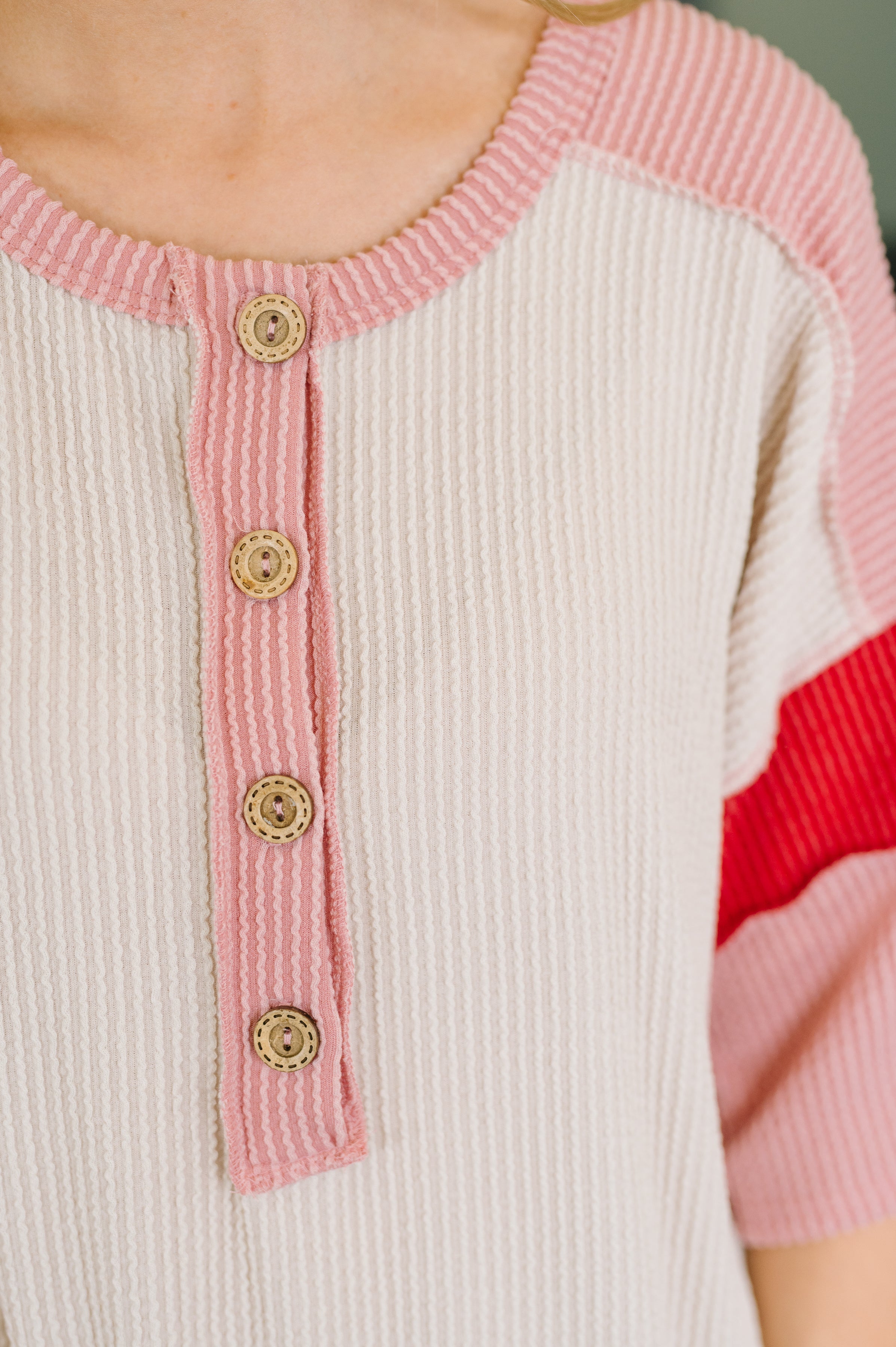 Close-up of cream top with pink ribbed round neckline and button placket, featuring pink and red contrast sleeve details