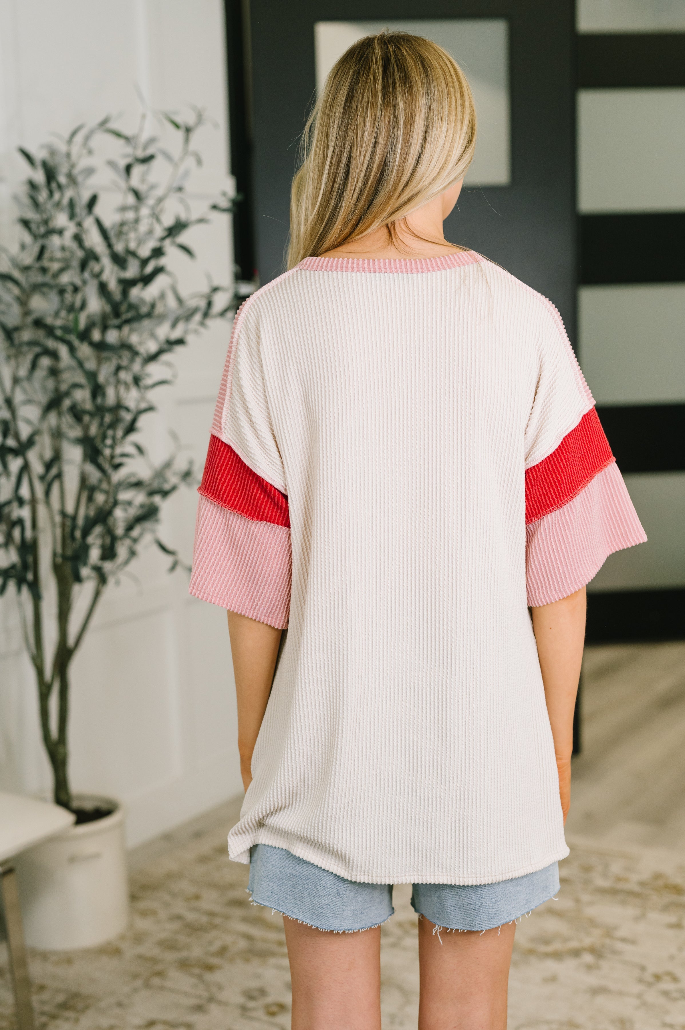 Back view of woman wearing a cream ribbed top with pink and red wide sleeves paired with light denim shorts indoors