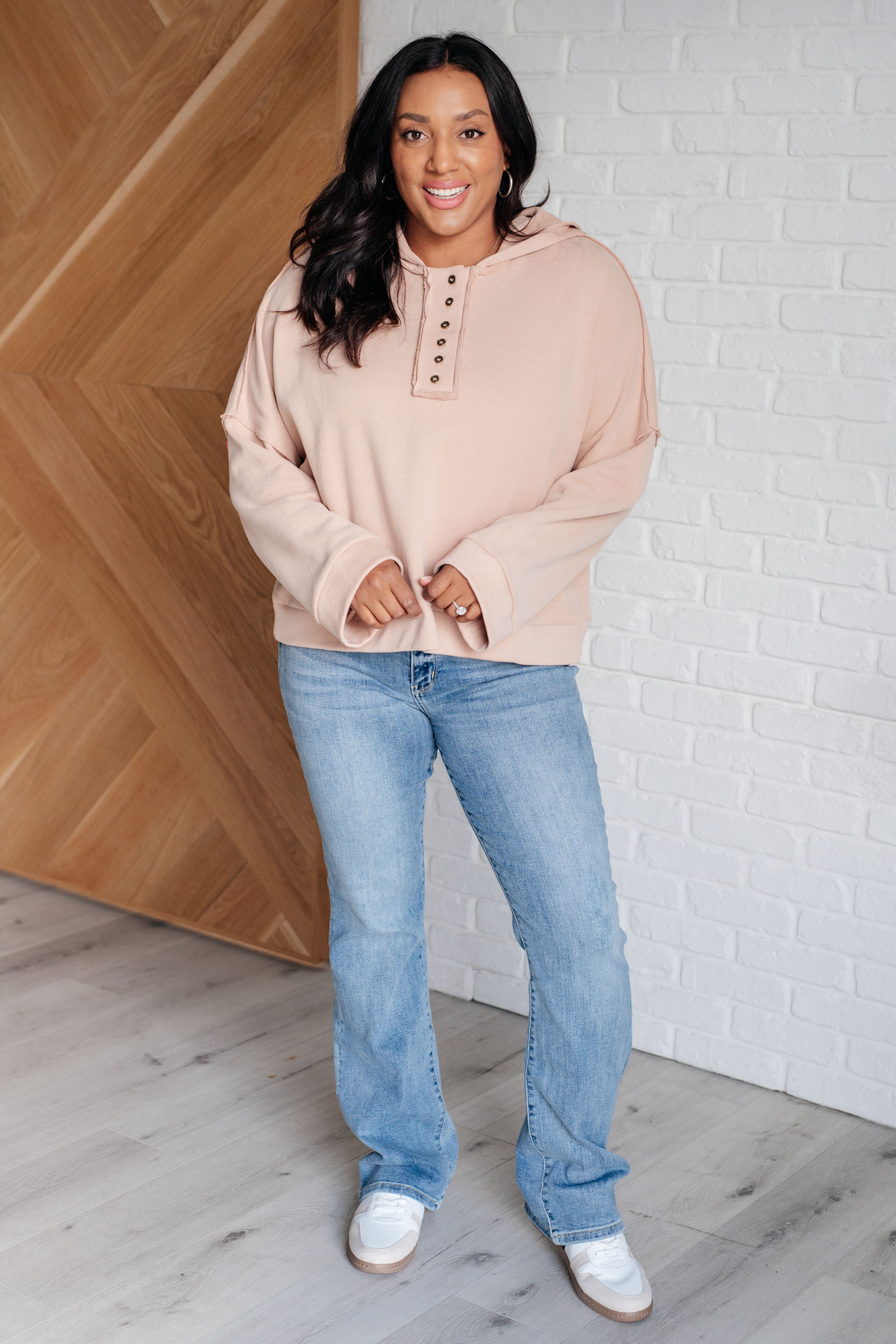 Woman wearing a beige cozy hoodie with button details paired with light blue jeans and white sneakers indoors