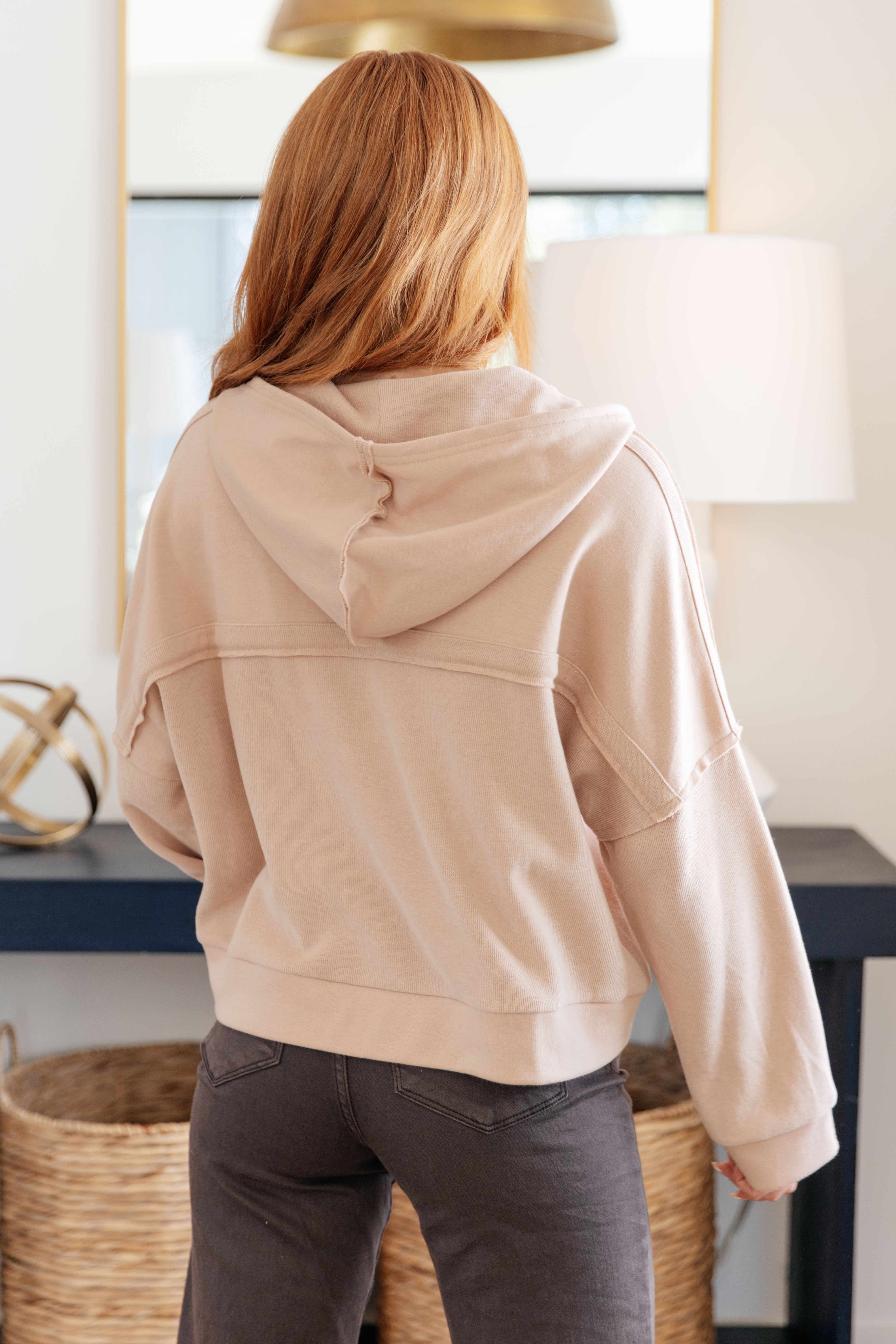 Woman with red hair wearing a beige oversized hoodie paired with dark jeans in a cozy indoor setting