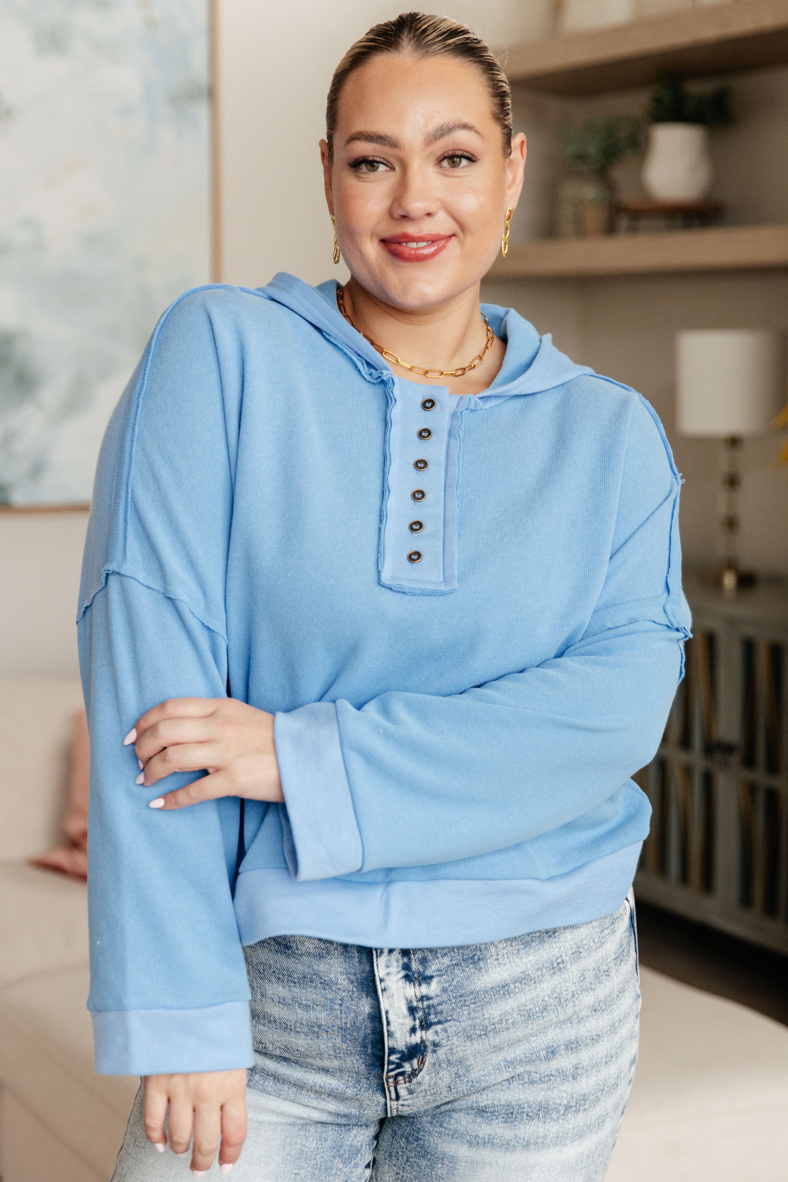 Plus-size woman wearing a sky-blue relaxed-fit hoodie with button details paired with light-wash jeans indoors smiling
