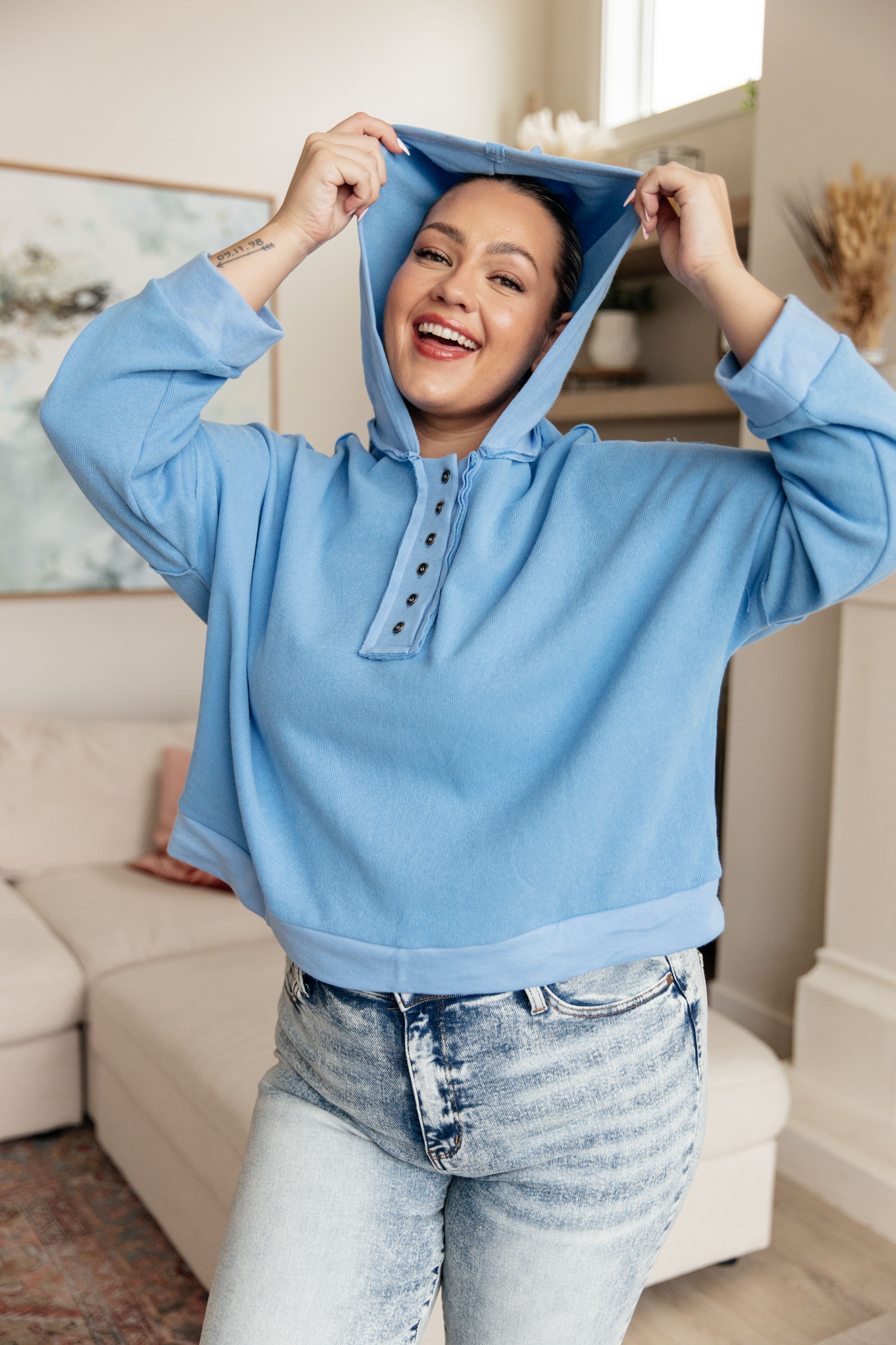 Smiling woman wearing a sky blue cropped hoodie with button details paired with light wash jeans indoors