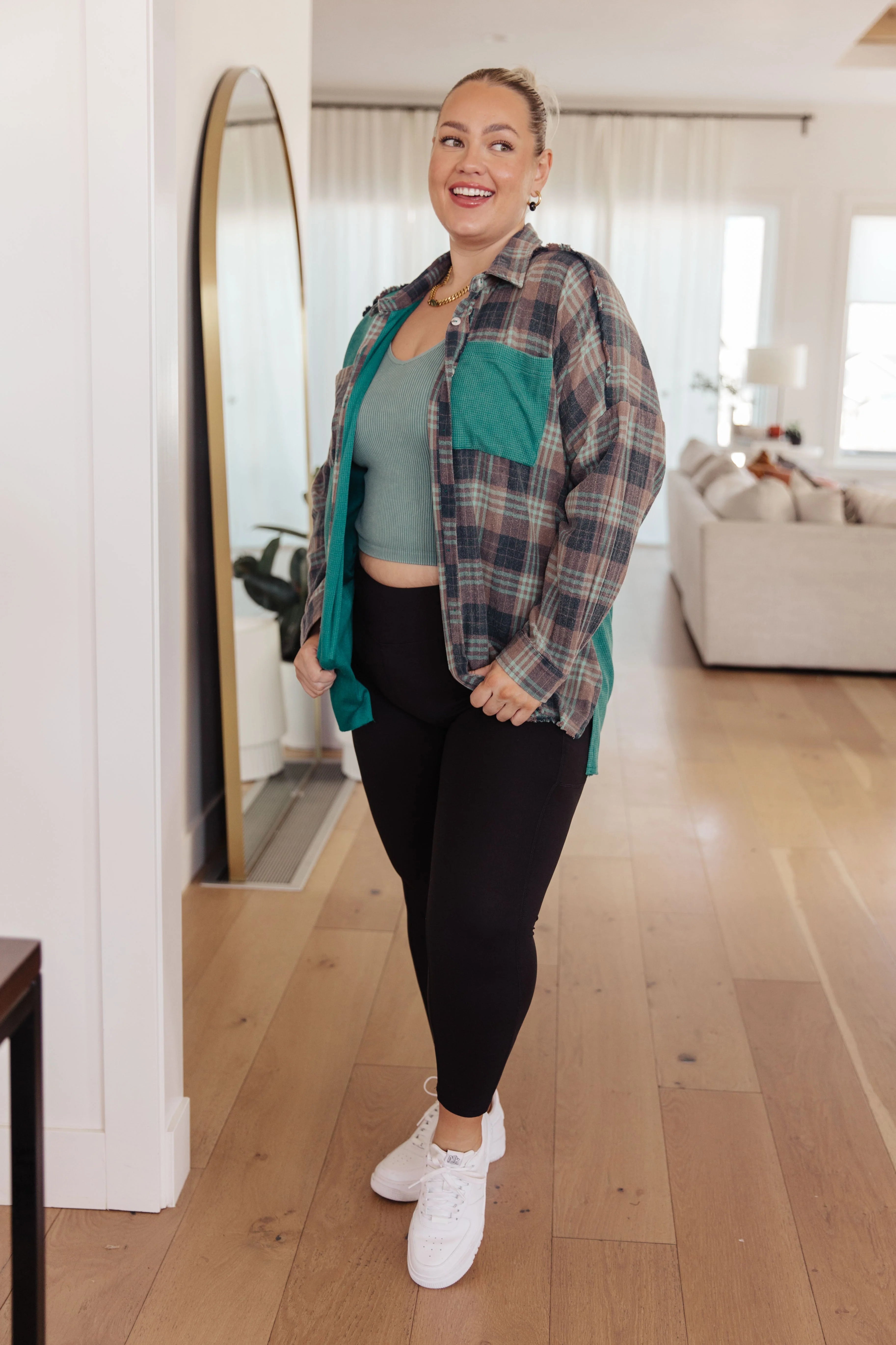 Curvy woman wearing a teal and brown plaid thermal shirt with black leggings and white sneakers indoors smiling