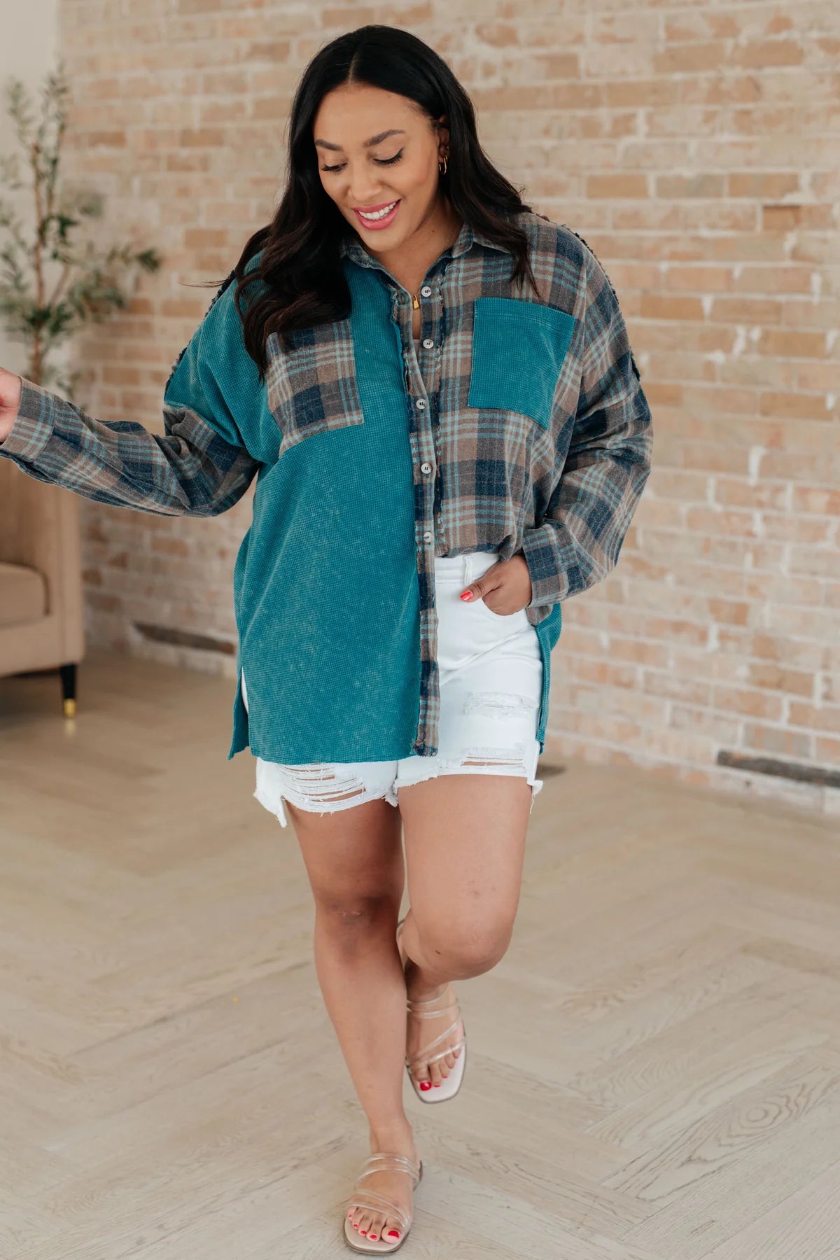 Woman wearing a teal plaid thermal button-up with white distressed shorts and sandals indoors smiling