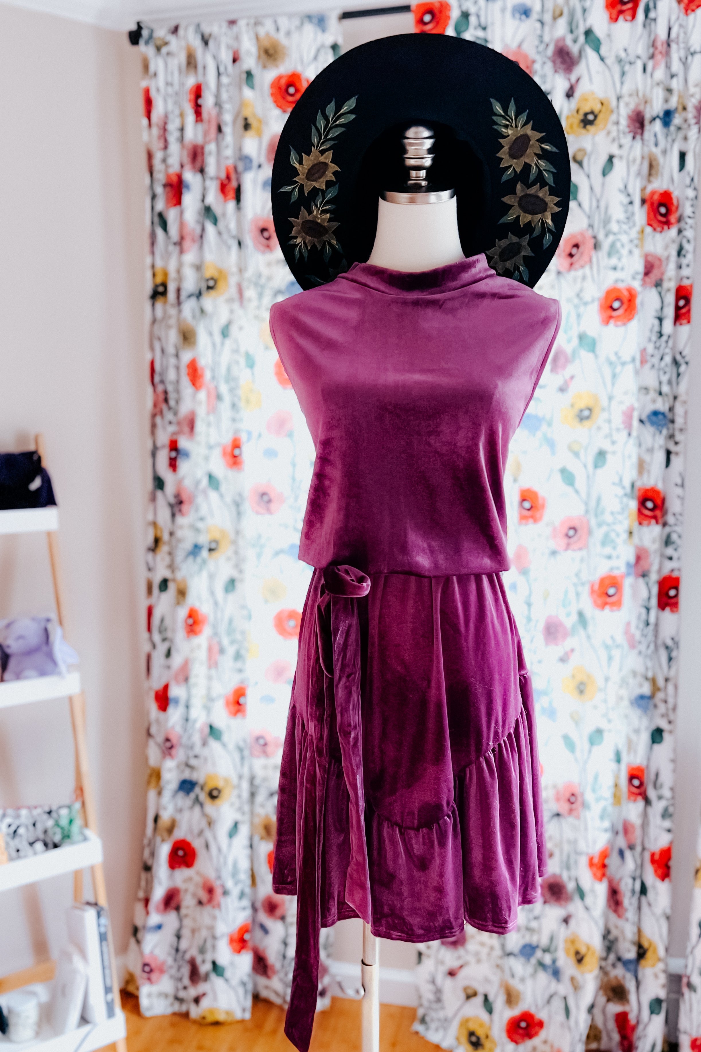 Purple velvet dress with a tied waist displayed on a mannequin wearing a black floral wide-brimmed hat indoors