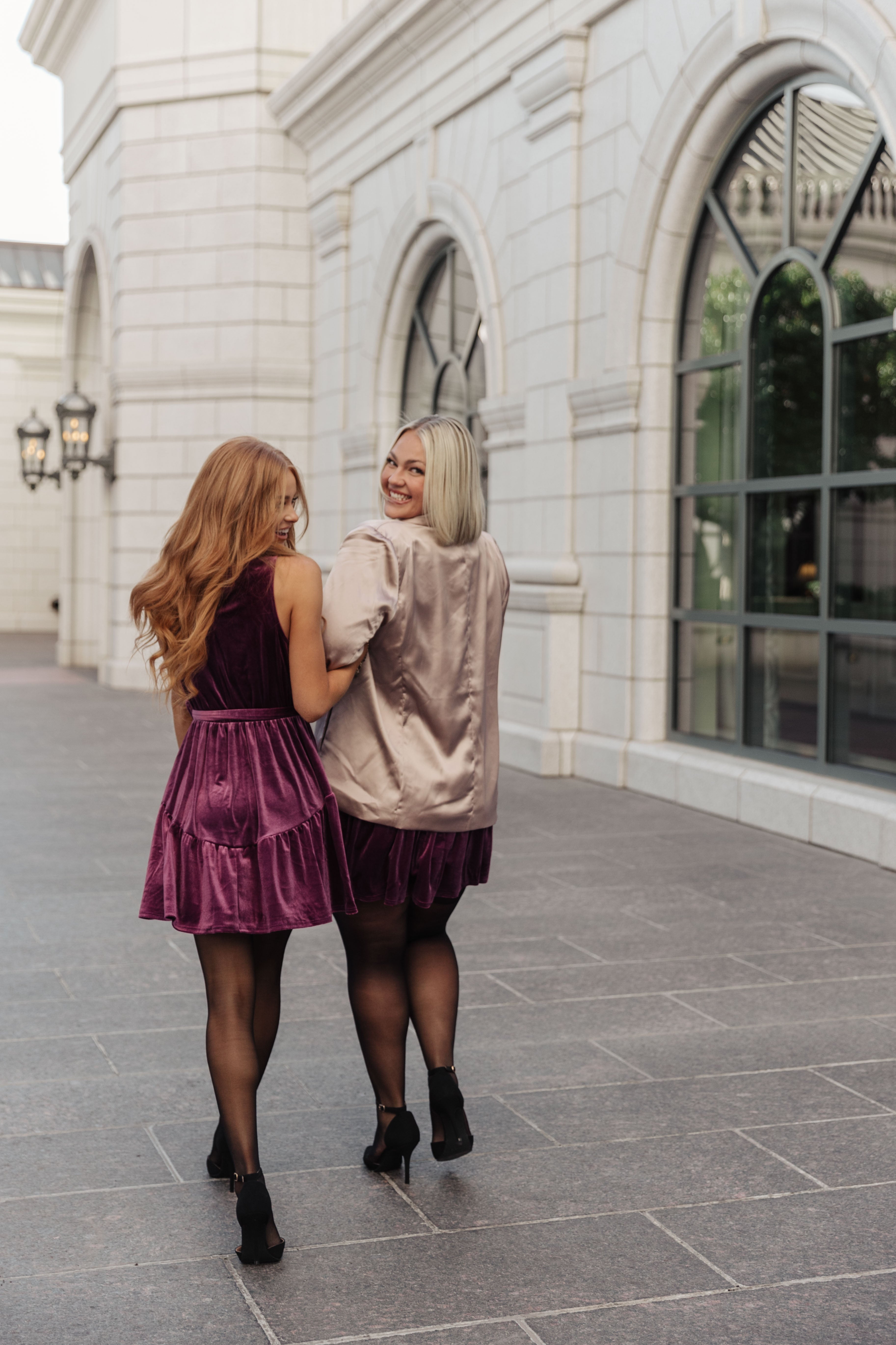 Two women walking arm in arm, one in a purple velvet bow dress and the other in a satin jacket with sheer black tights outdoors
