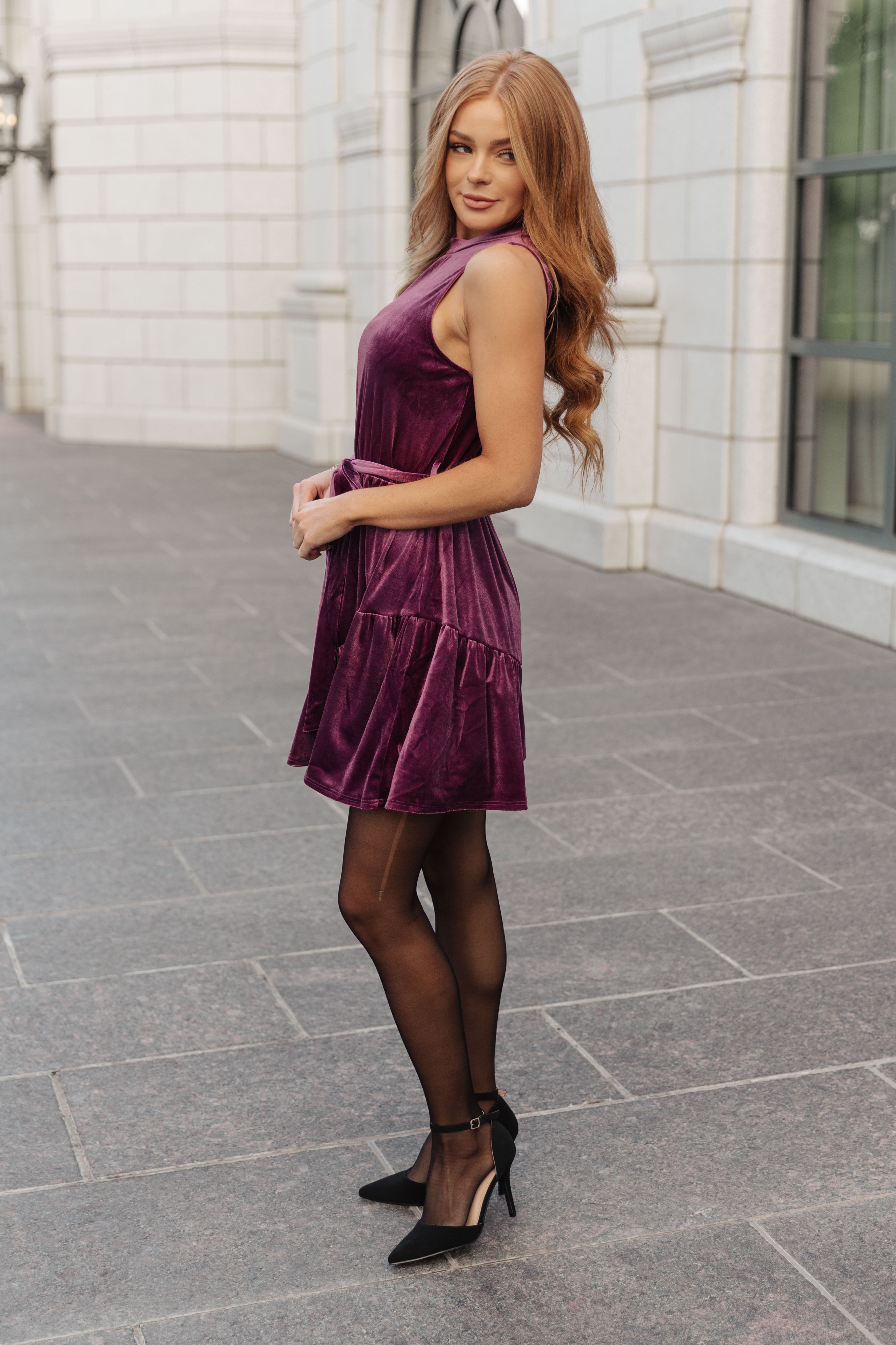 Woman wearing a purple velvet dress with side ties paired with sheer black tights and black heels outdoors