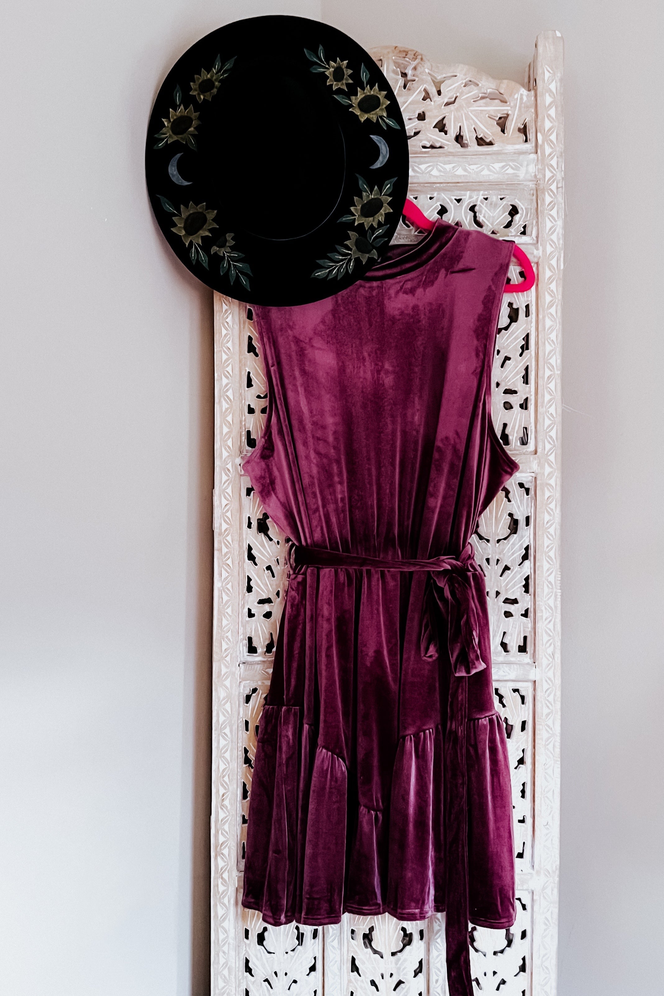 Purple velvet sleeveless dress with a tied waist hanging next to a black hat with floral and moon designs