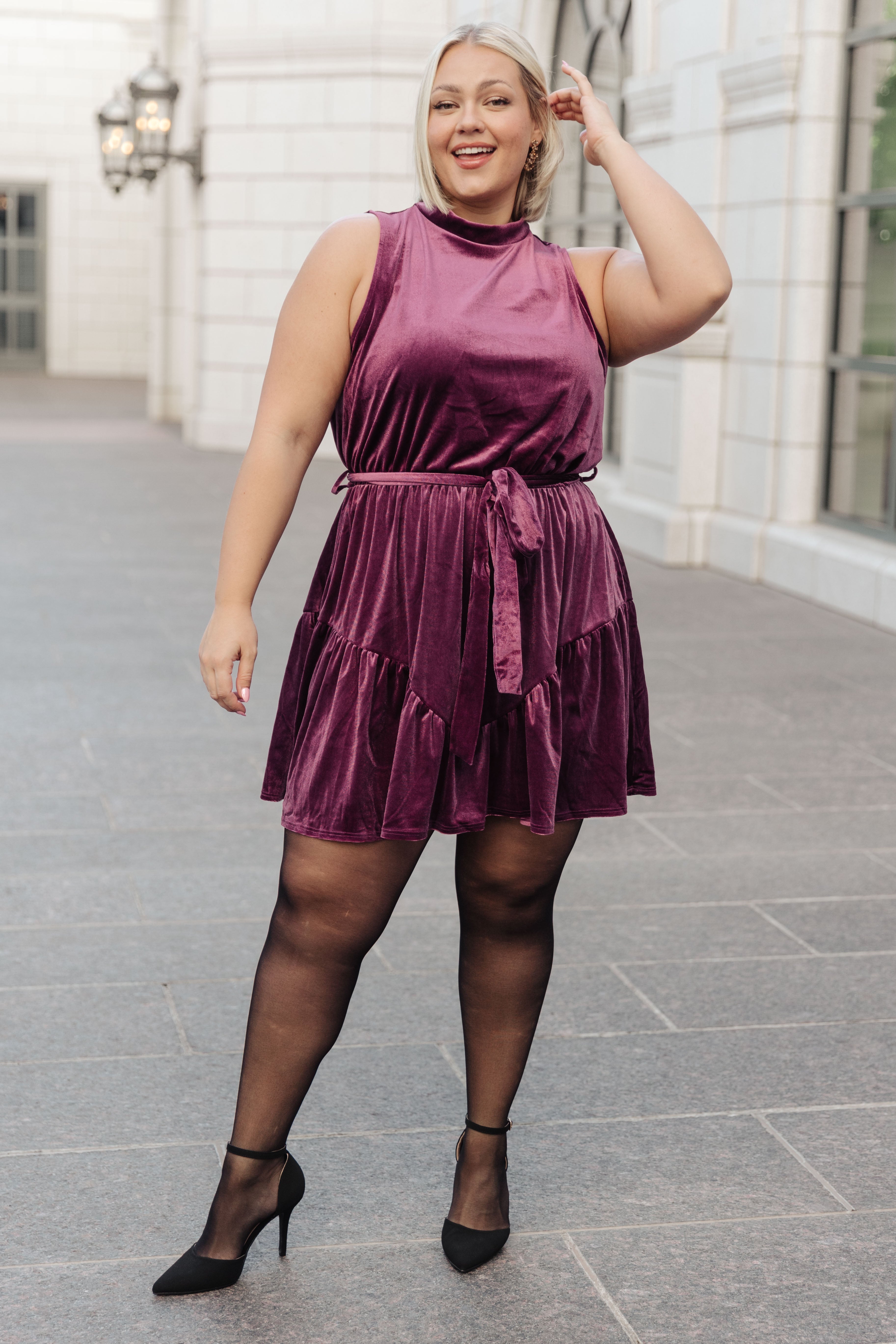 Plus-size woman wearing a sleeveless purple velvet dress with a waist tie and black sheer tights, outdoors smiling confidently