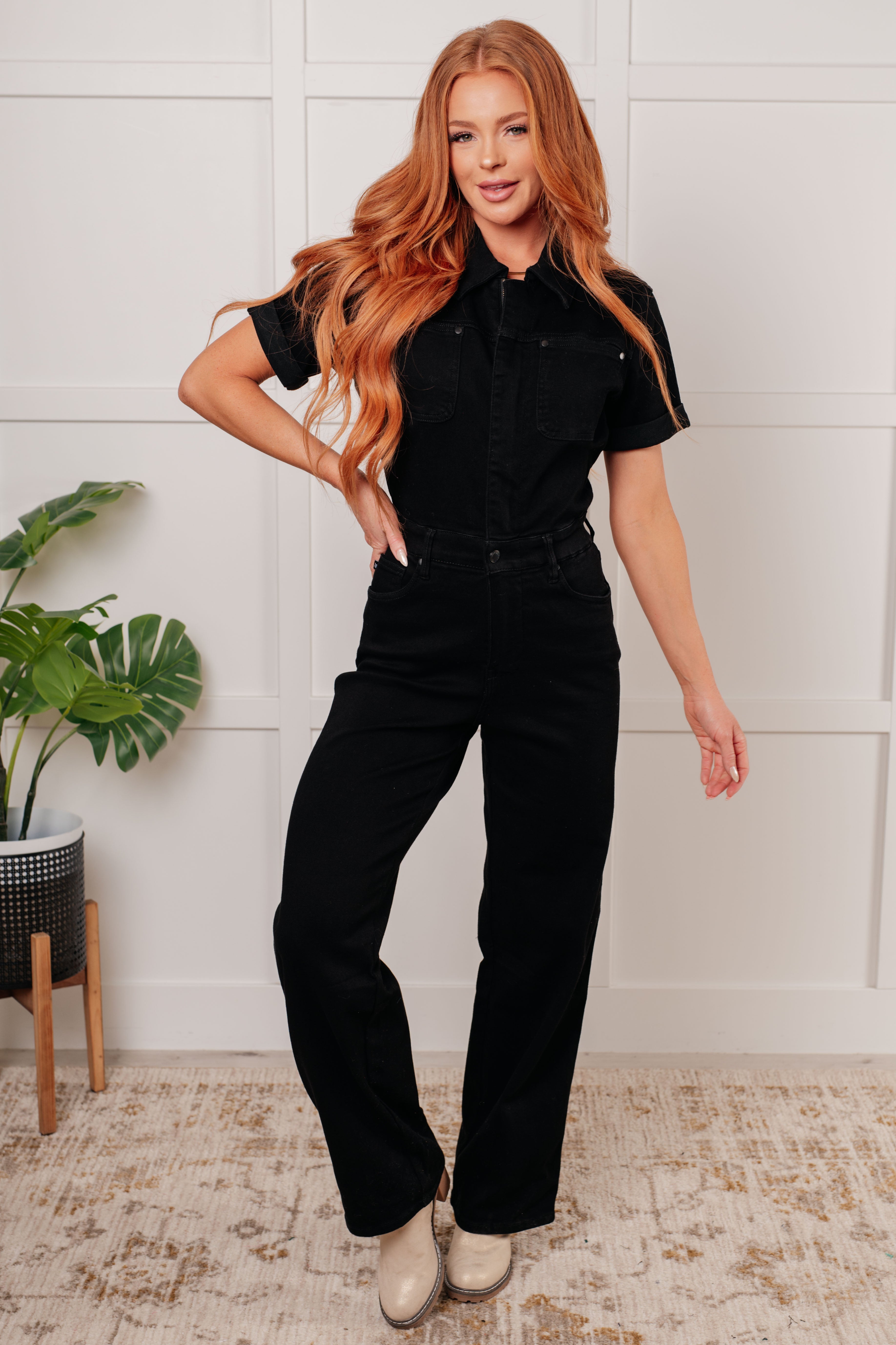 Woman standing indoors wearing black short sleeve denim jumpsuit with collar and front pockets