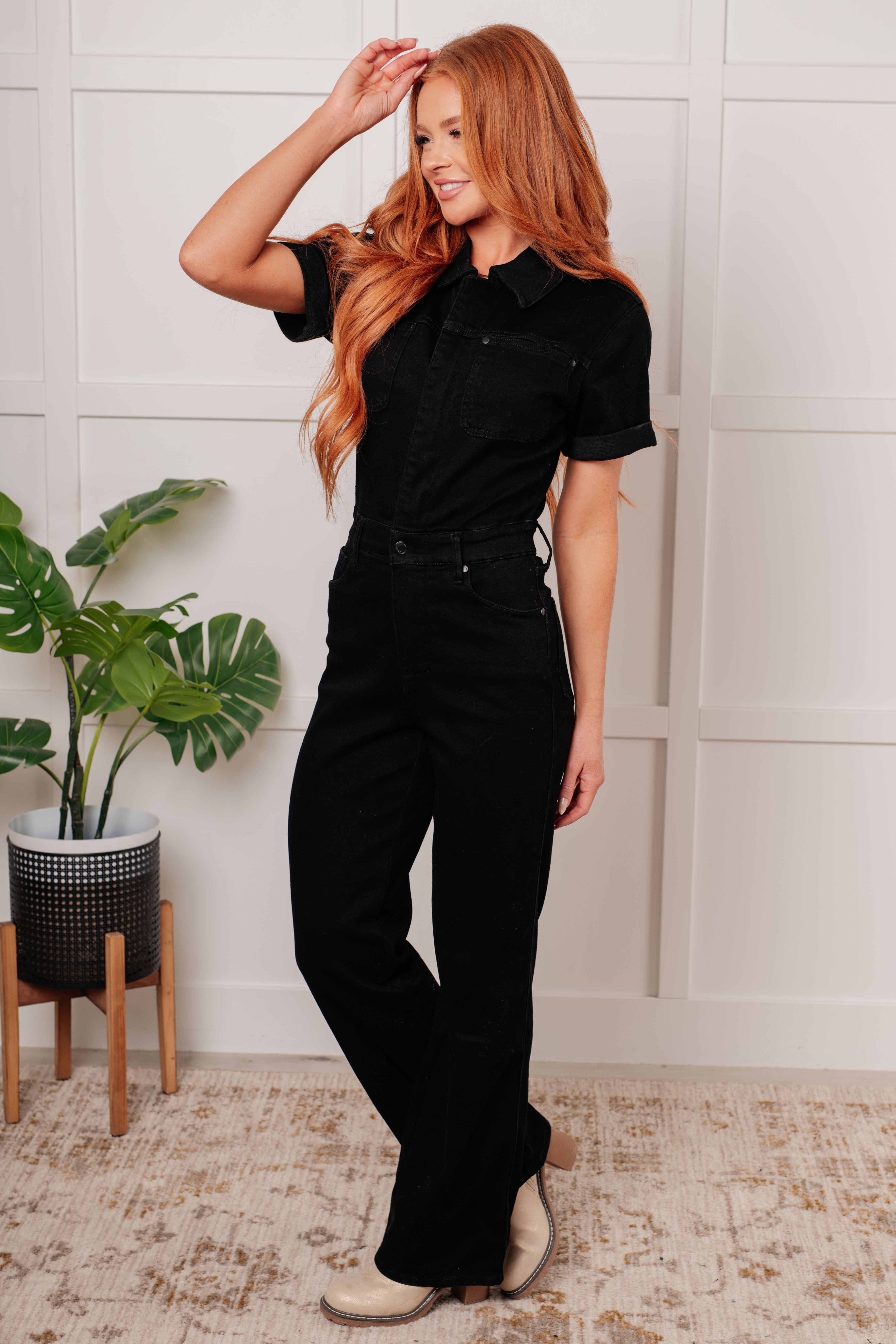 Woman smiling wearing black short sleeve denim jumpsuit with collar standing indoors near plant