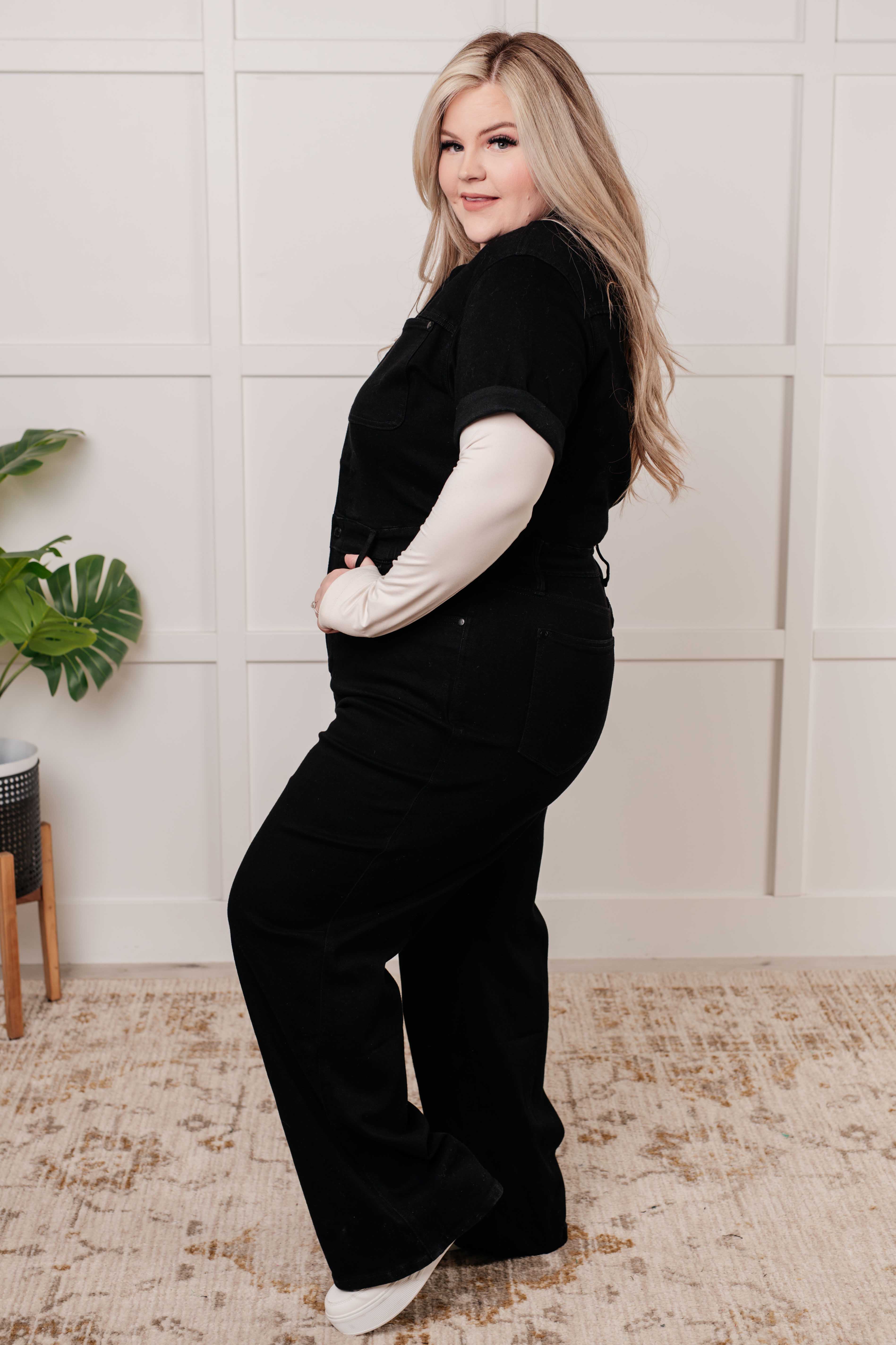Side view of woman wearing black short sleeve control top denim jumpsuit with light long sleeve underlayer indoors