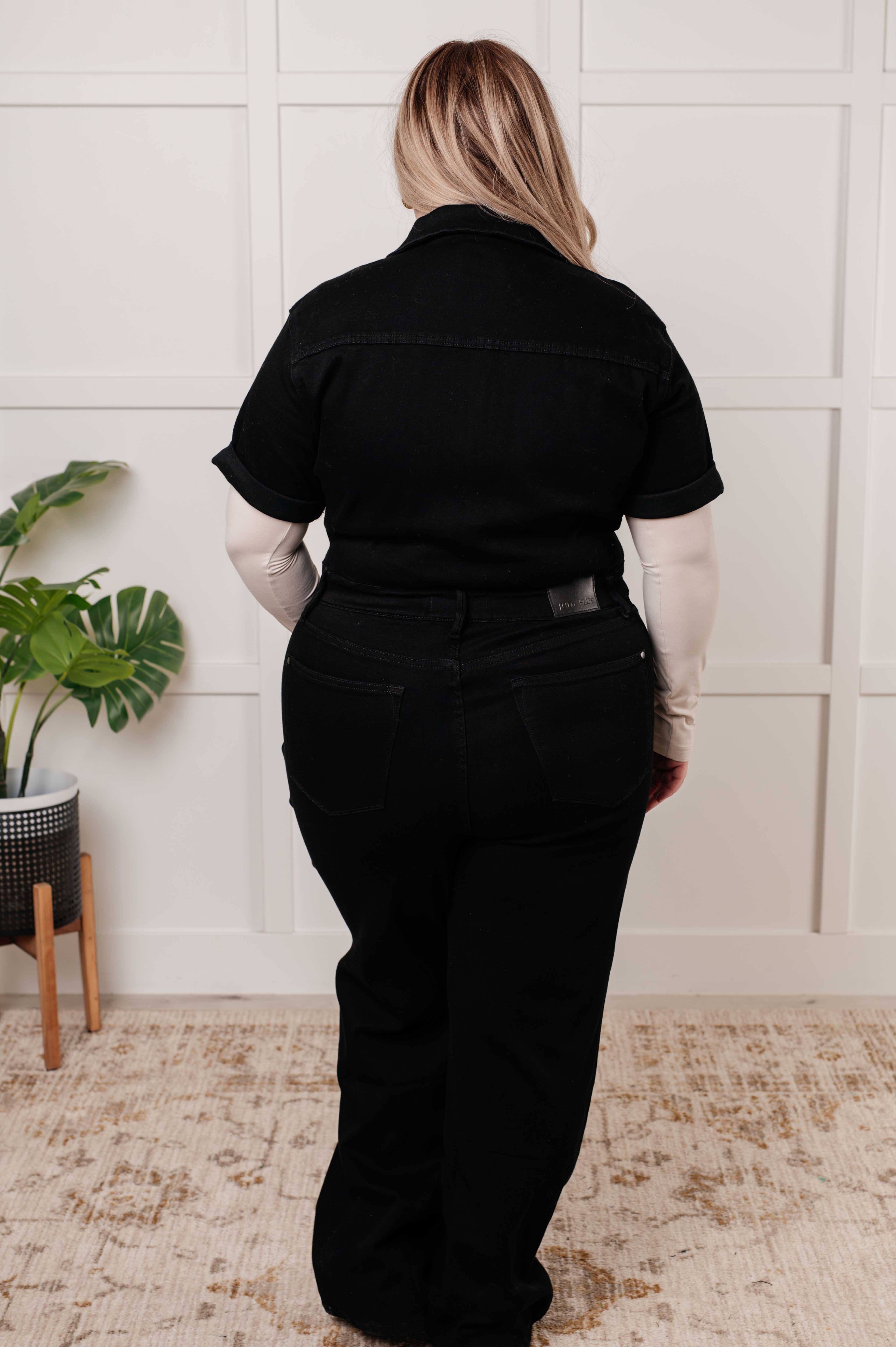 Back view of woman wearing black short sleeve control top denim jumpsuit indoors with beige long sleeve underlayer
