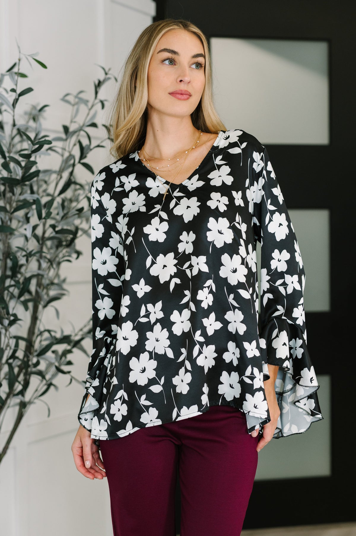 Woman wearing a black floral blouse with a V neckline and flowing ruffled sleeves paired with burgundy pants in a modern room