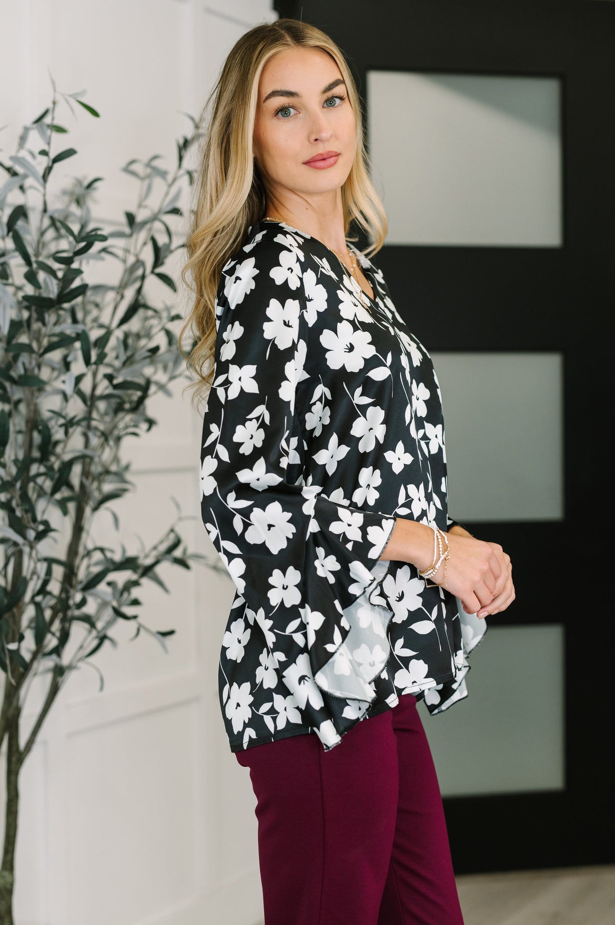 Woman wearing a black floral blouse with flowing ruffled sleeves and burgundy pants in a modern entryway