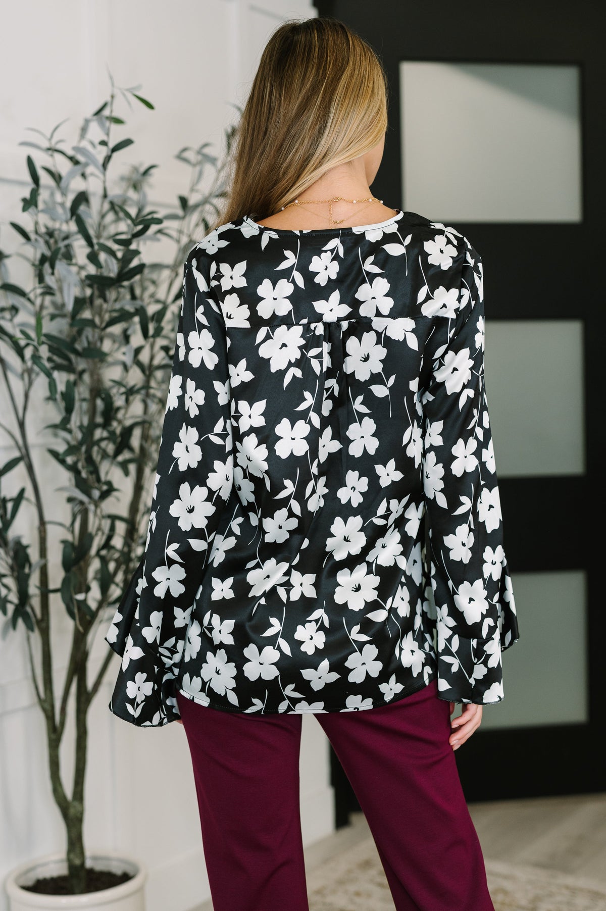 Back view of a woman wearing a black floral blouse with ruffled sleeves and burgundy pants in a modern interior