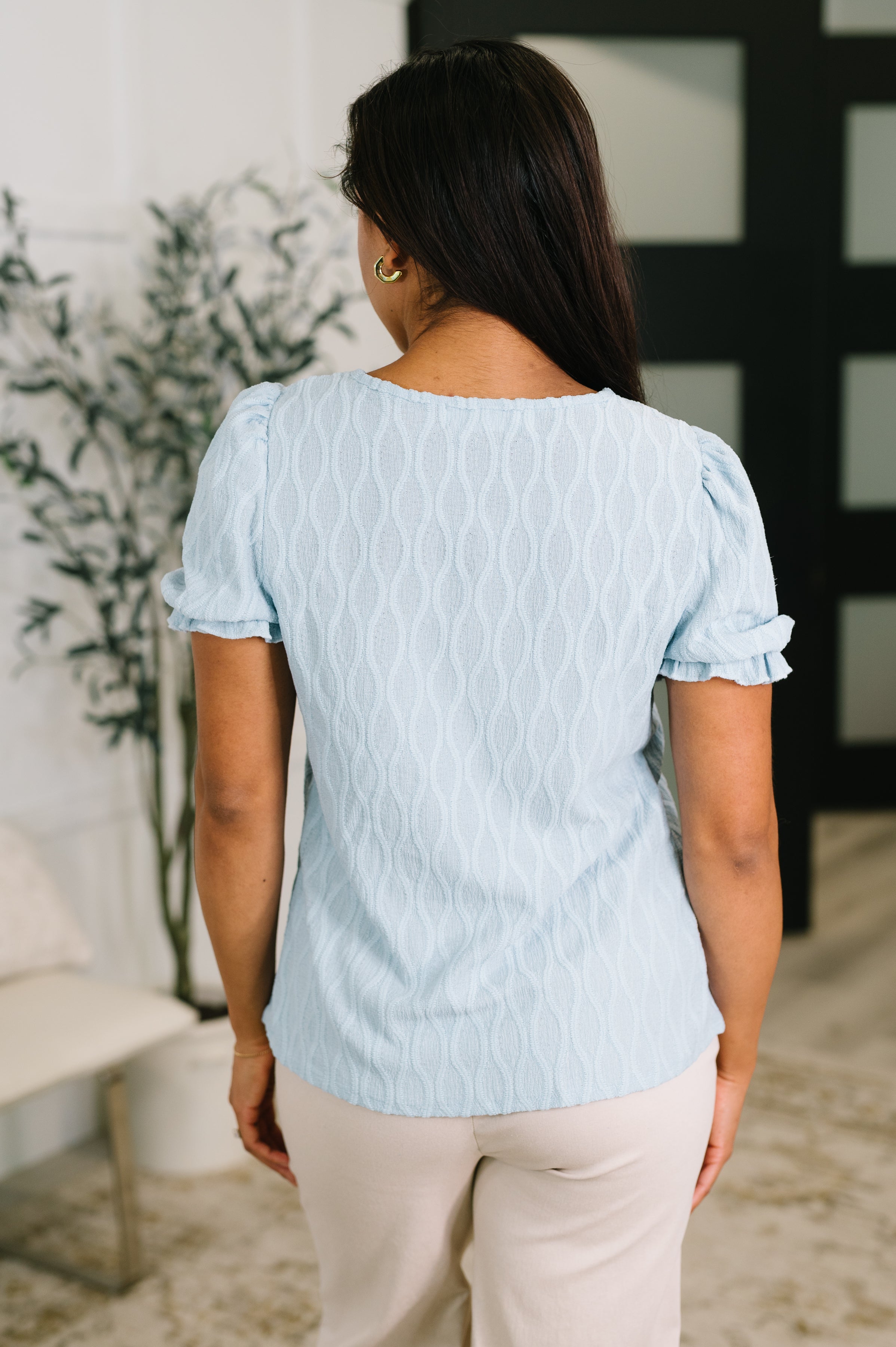Woman wearing powder blue textured blouse with short puff sleeves standing indoors with back to camera