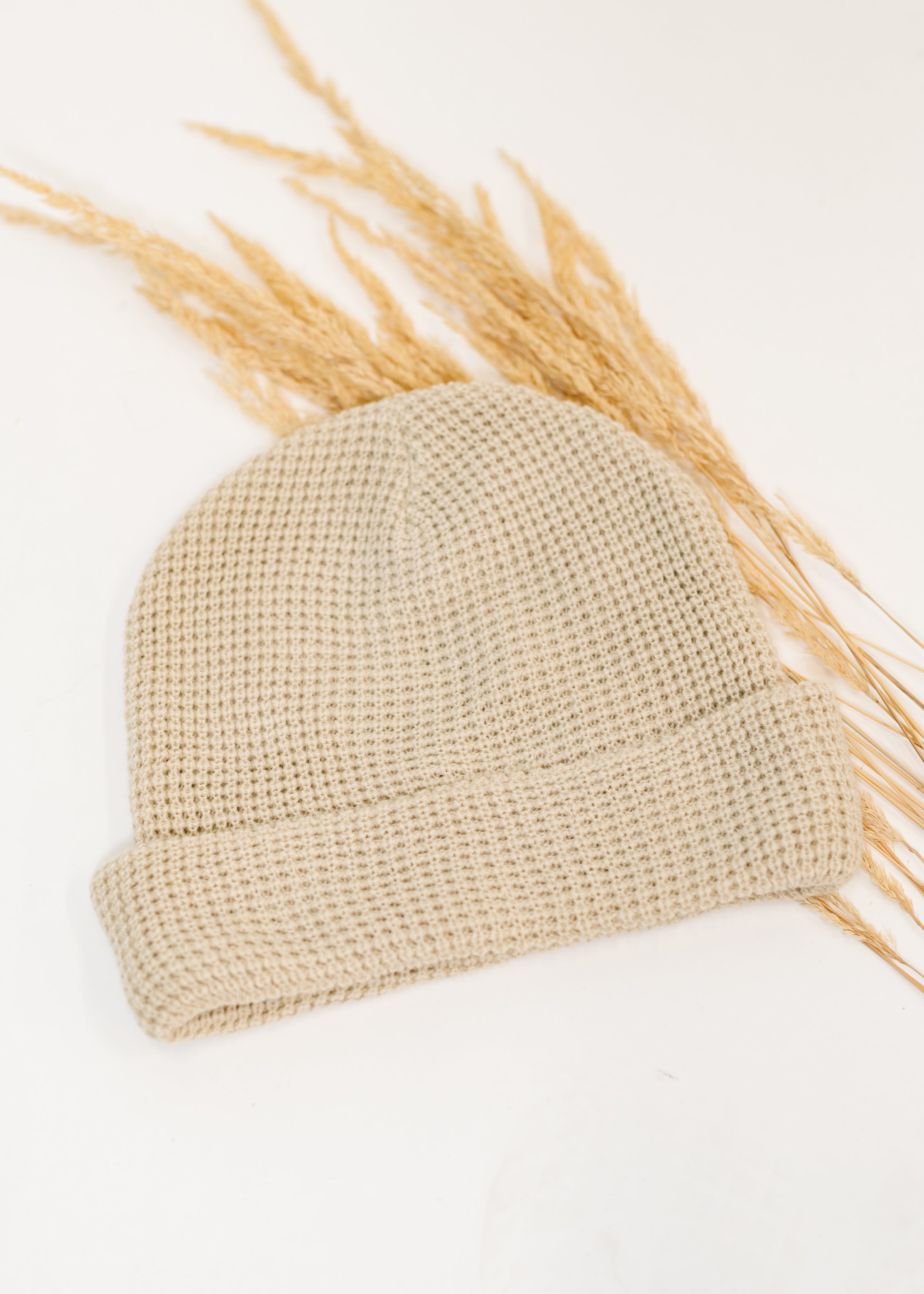 Beige waffle knit beanie with folded cuff laid on white surface alongside dried grass accents