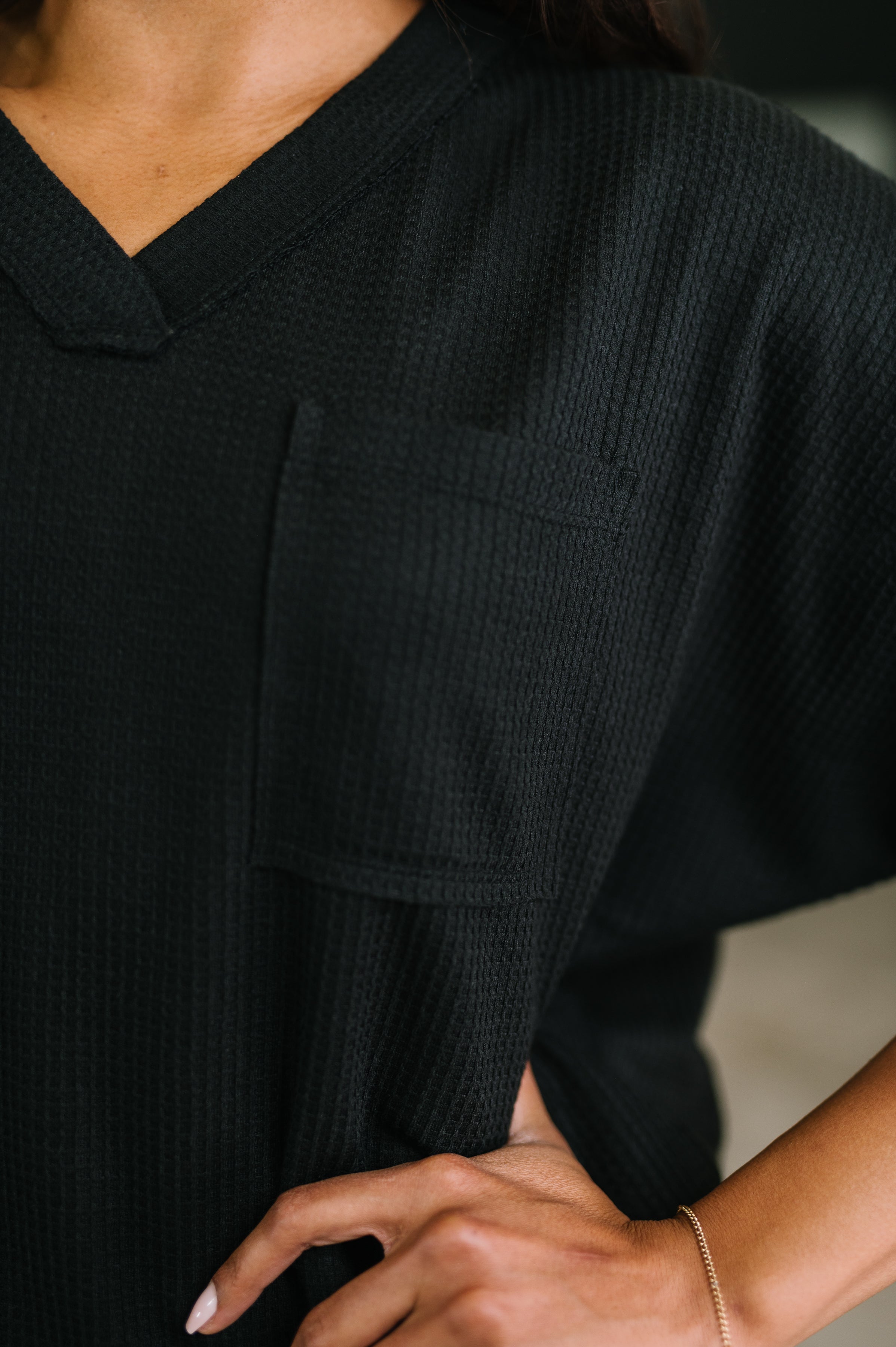 Close-up of a black textured V-neck top with chest pocket worn by a person with hand on hip indoors