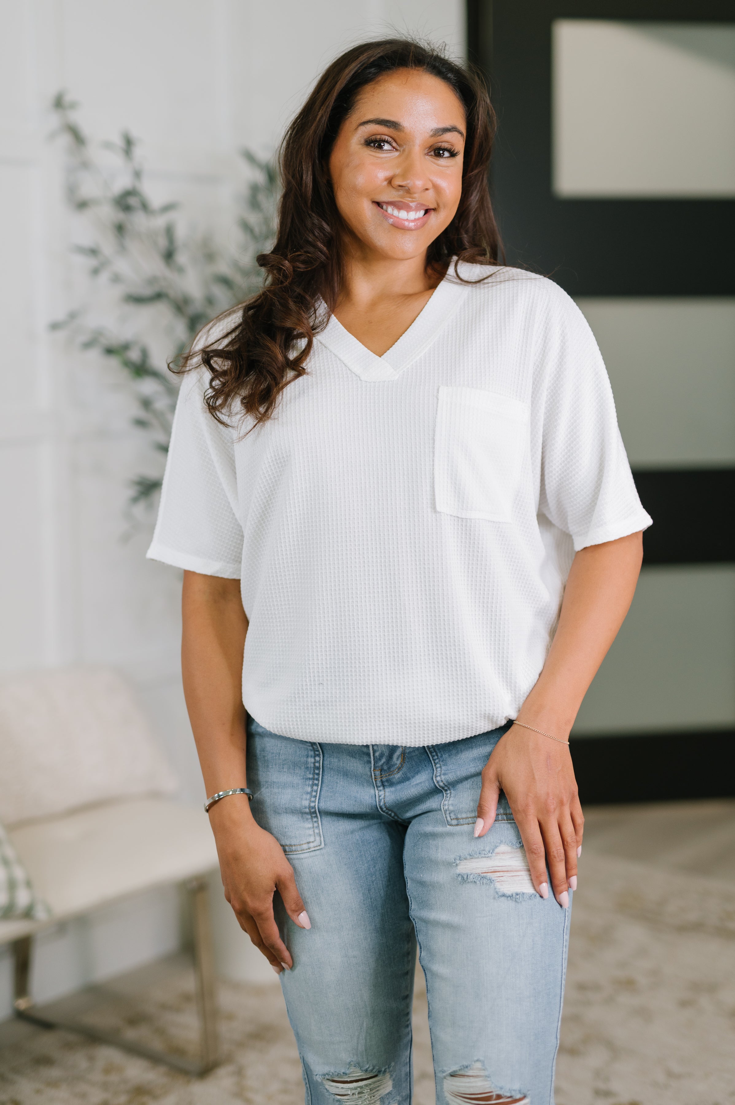 Smiling woman wearing an ivory textured V-neck tee with chest pocket and distressed light blue jeans indoors