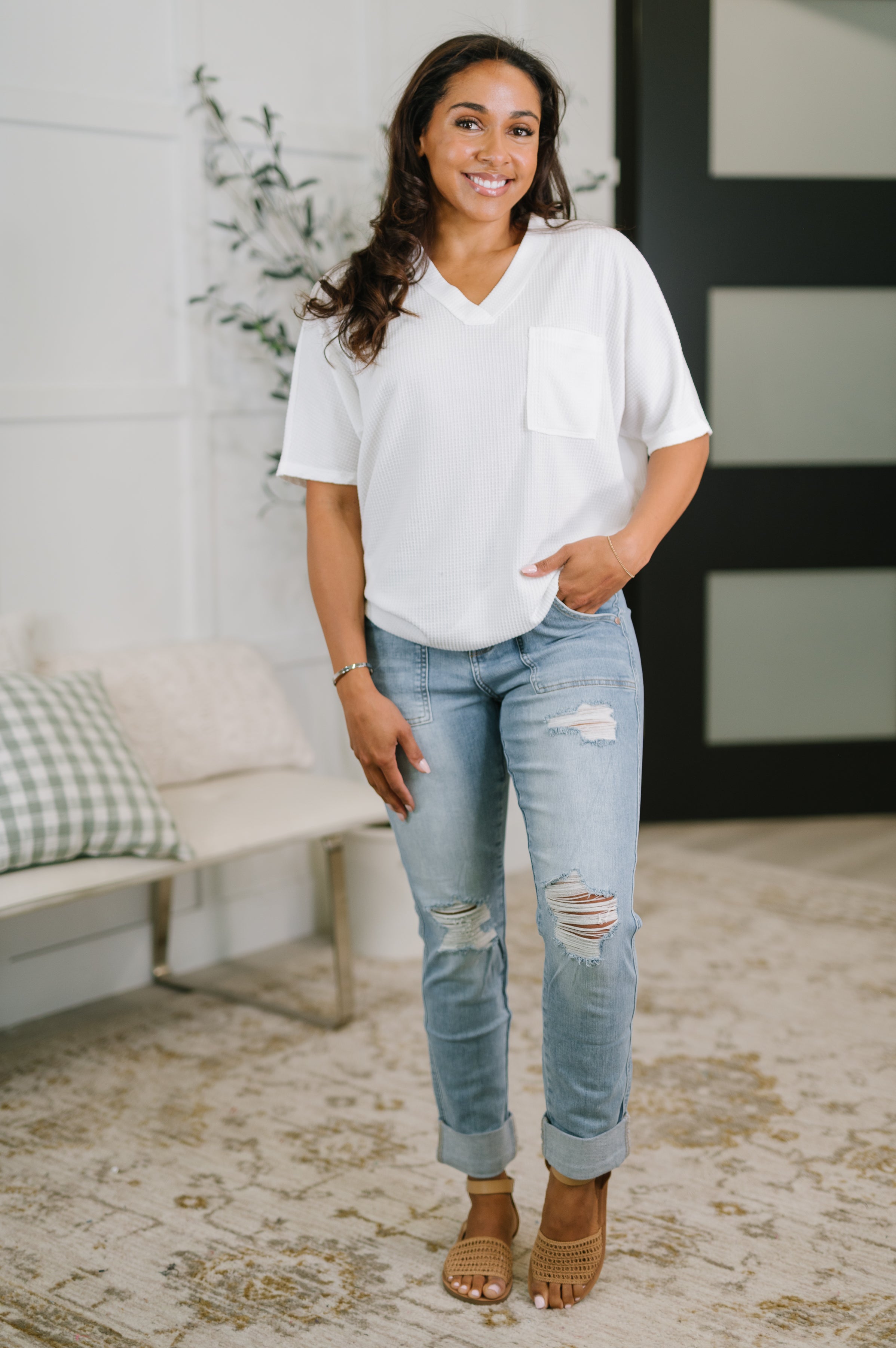 Smiling woman wearing an ivory relaxed v-neck tee with pocket and ripped light wash jeans in a cozy indoor setting