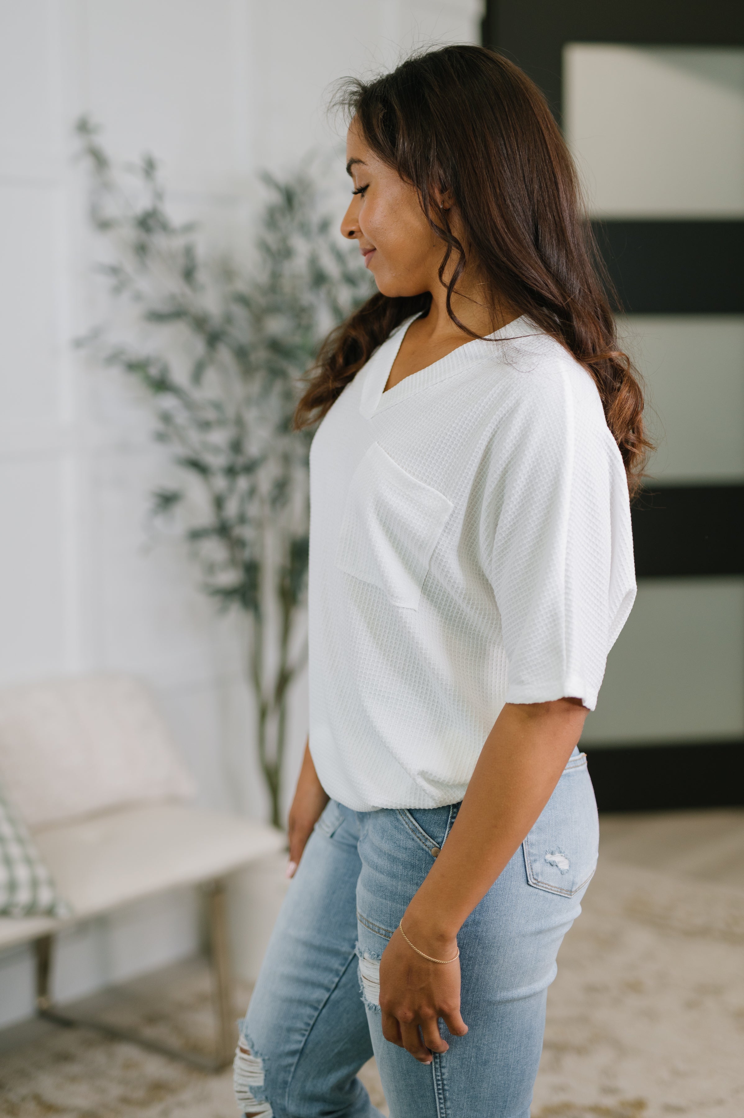 Woman wearing ivory textured v-neck tee with pocket and light ripped jeans standing indoors next to plant and chair