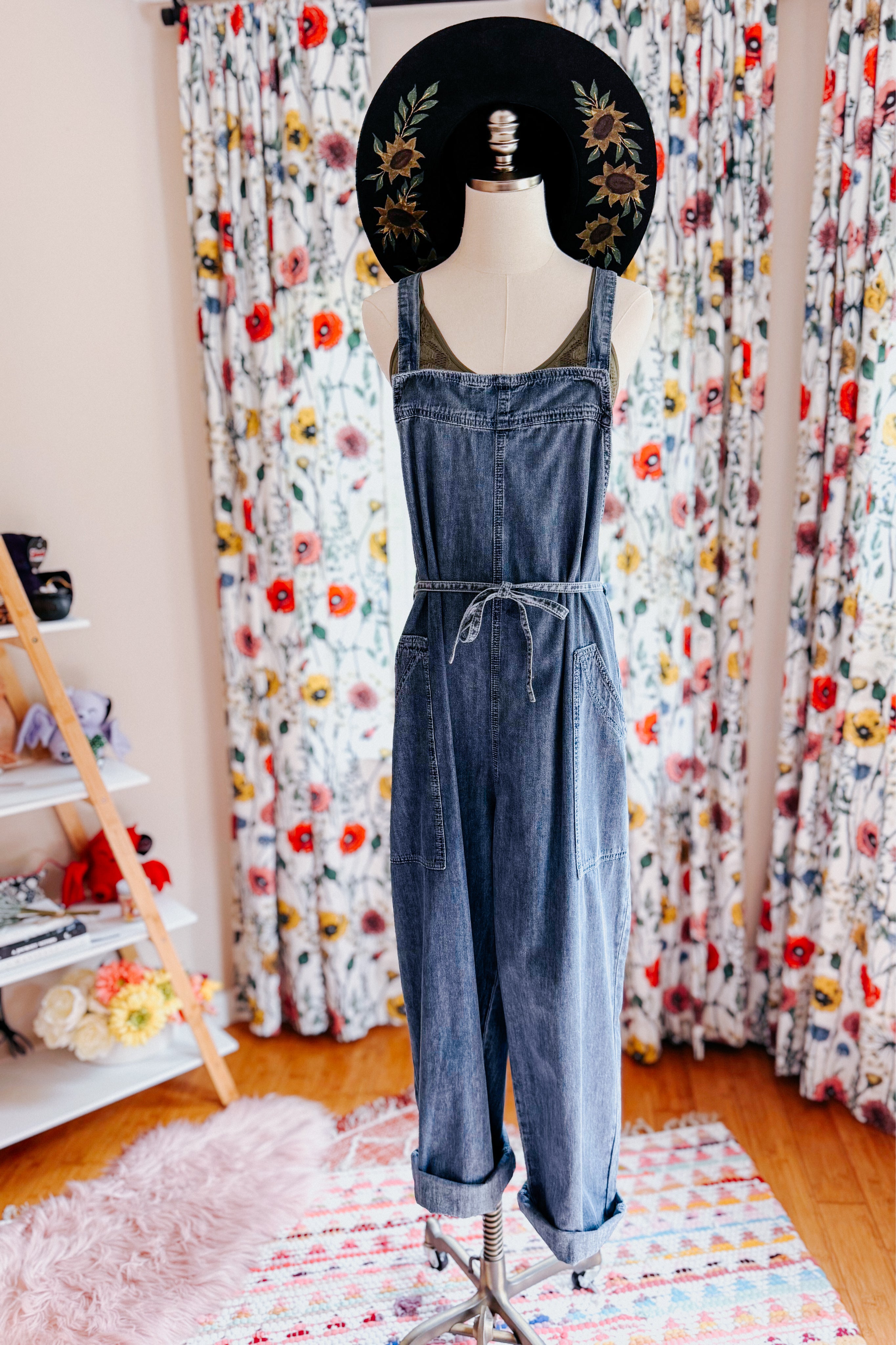 Dark blue denim waist tie overalls styled on mannequin with wide-brimmed black hat and floral curtain background