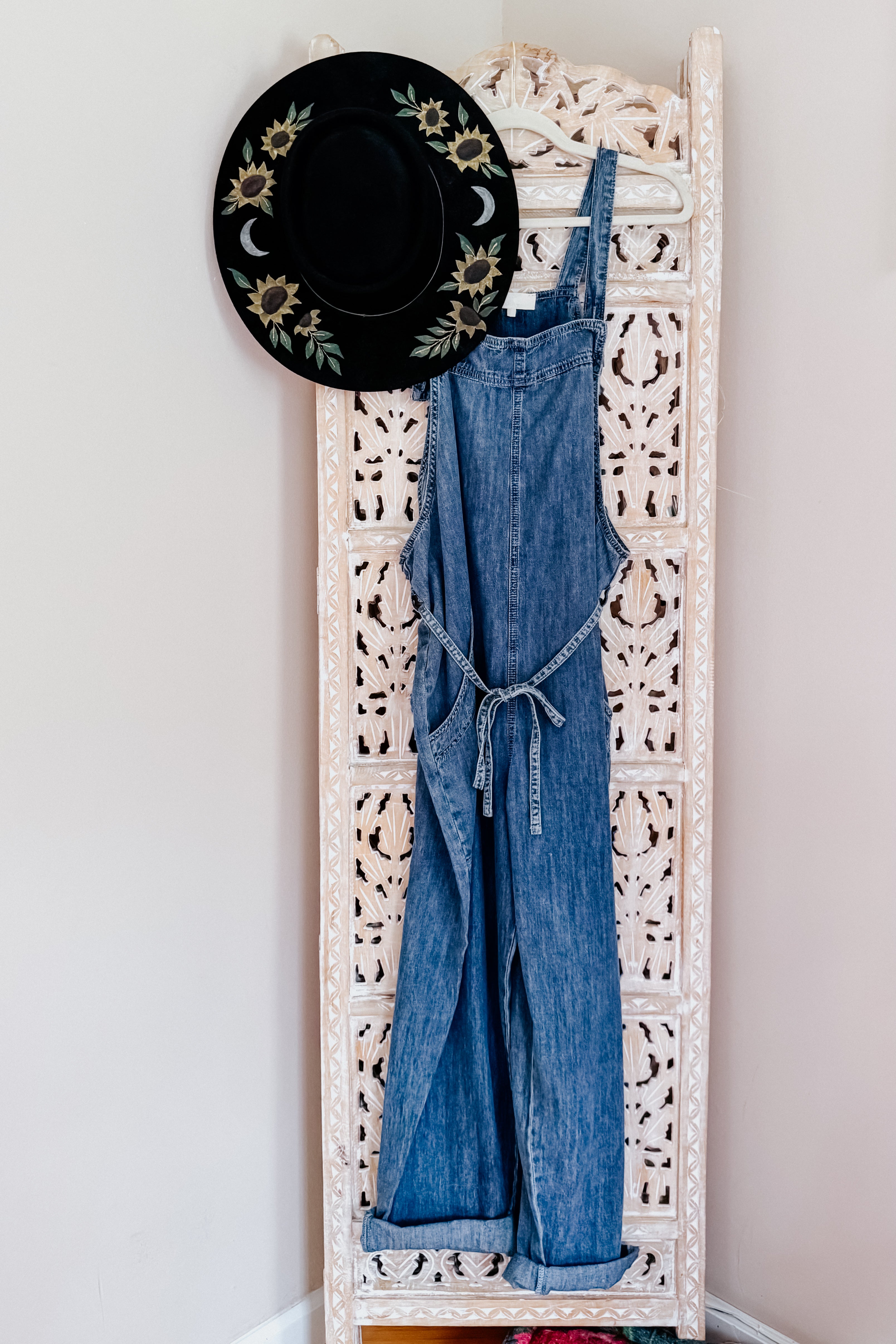 Dark blue denim waist tie overalls hanging on carved cream room divider with black floral hat on side