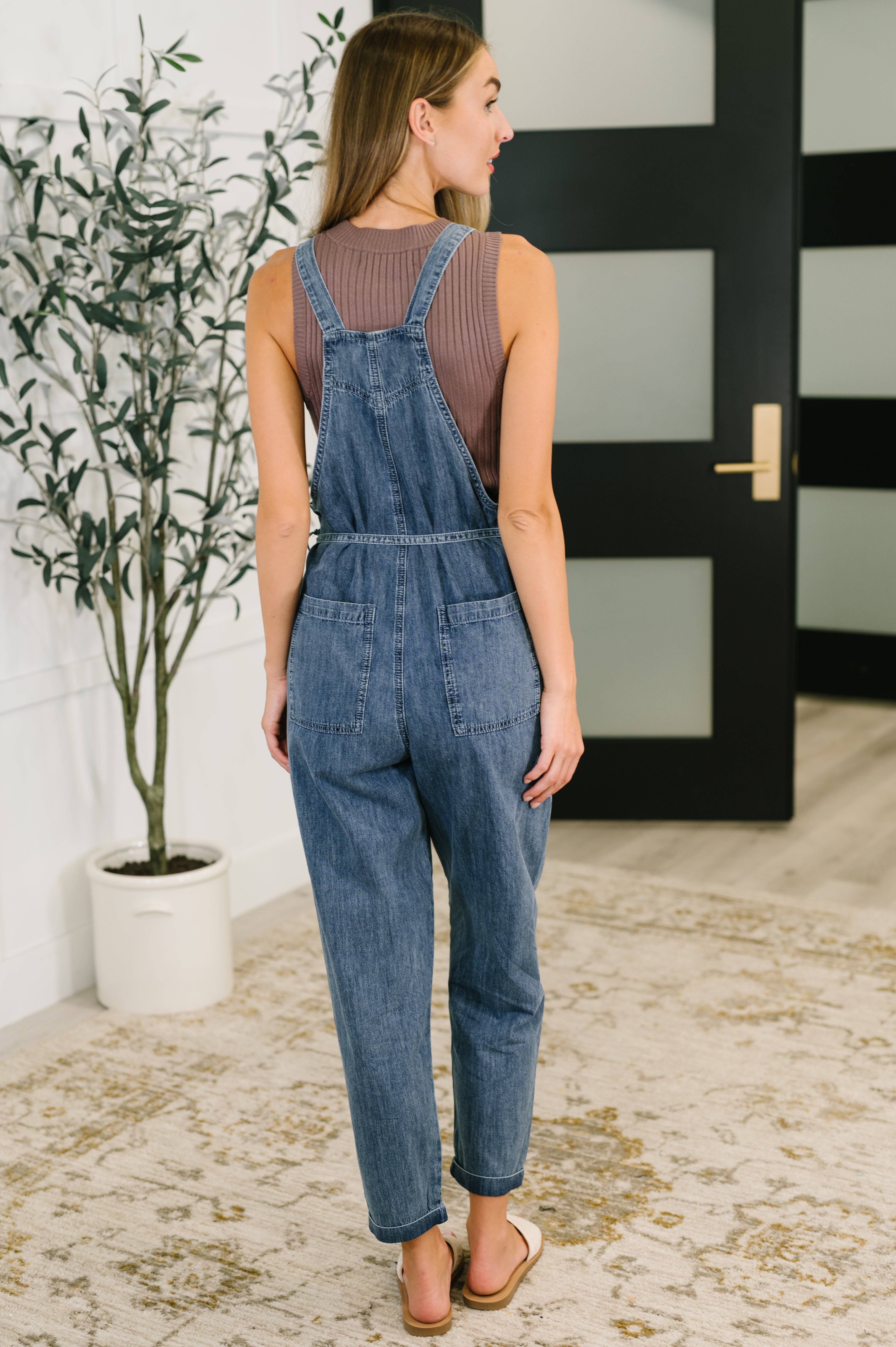 Woman wearing dark blue denim waist tie overalls with sleeveless ribbed brown top and sandals indoors