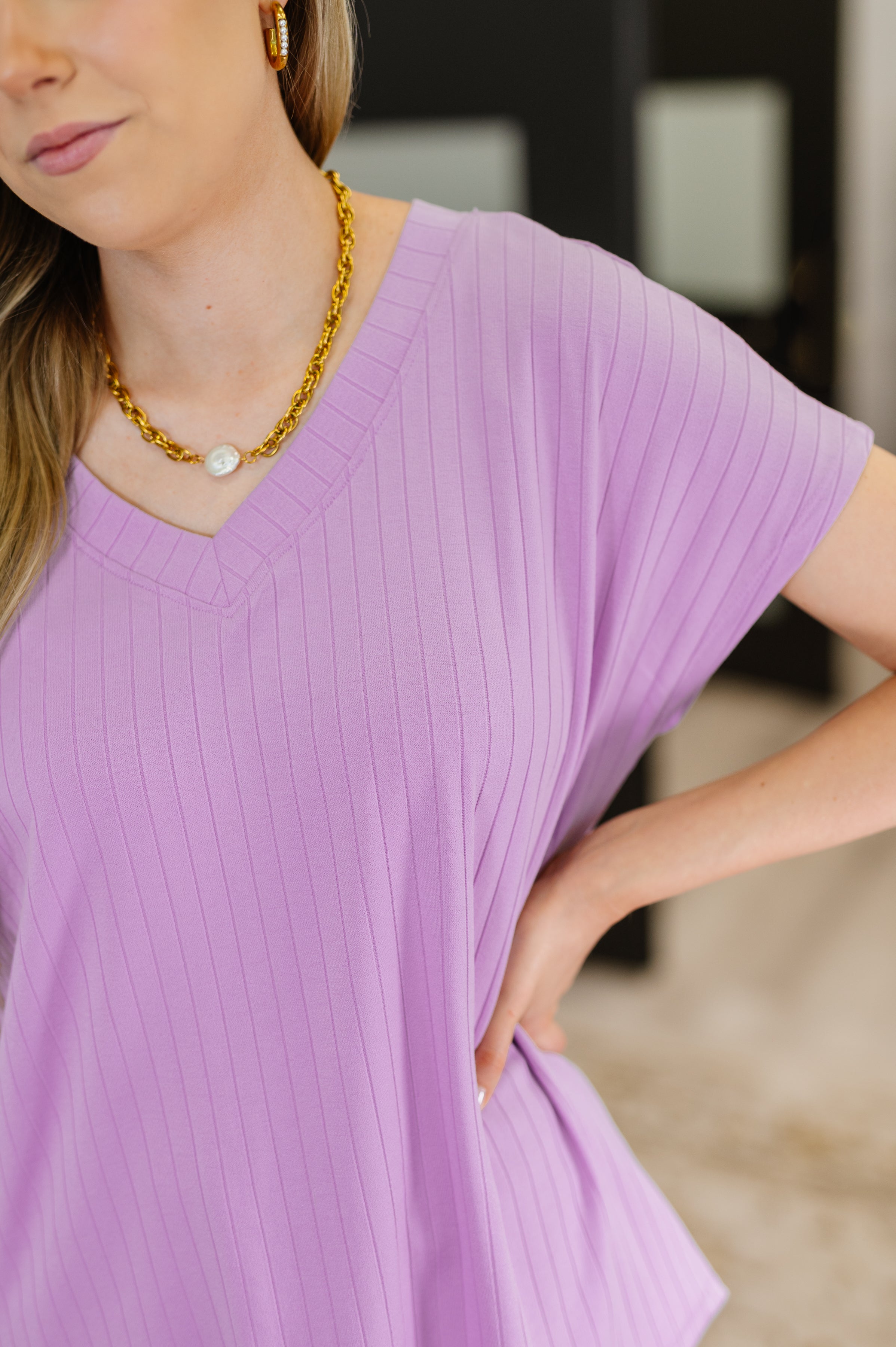 Close-up of a woman wearing a ribbed lavender V-neck top with short sleeves and gold chain necklace with a pearl pendant