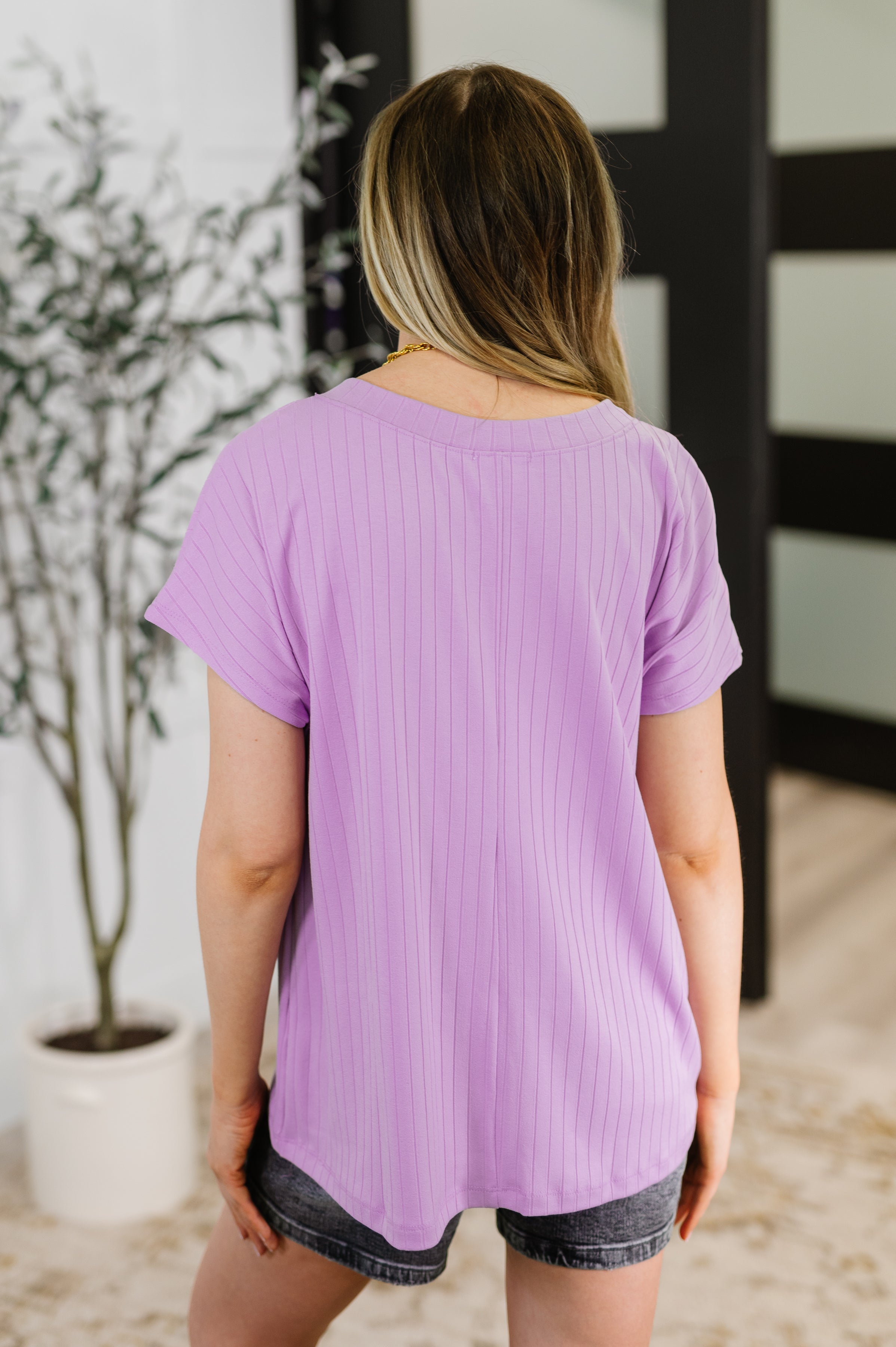 Woman seen from behind wearing a lavender ribbed short sleeve top paired with dark denim shorts