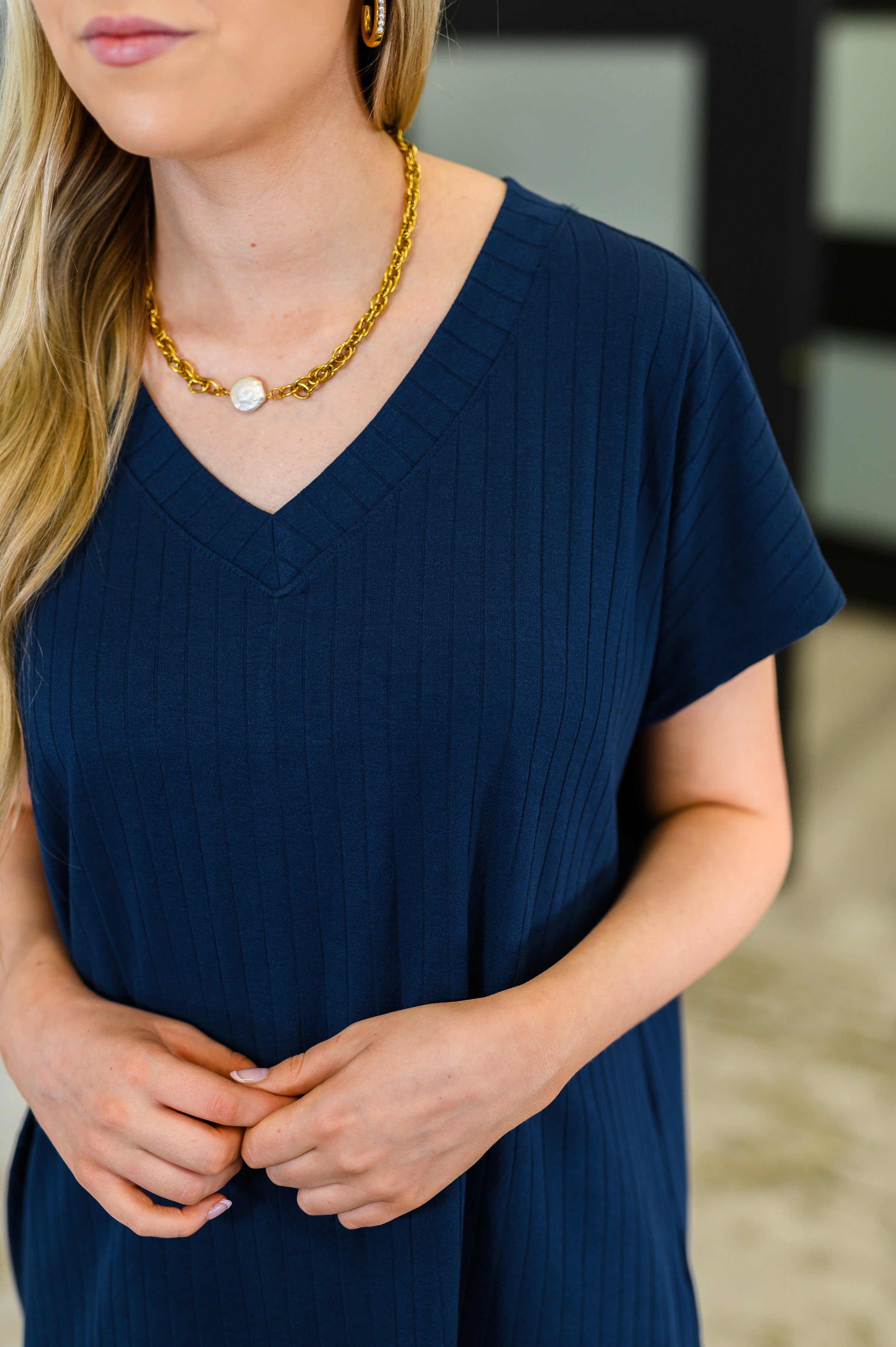 Woman wearing a navy ribbed V-neck tee with short sleeves and gold chain necklace featuring a white pendant indoors
