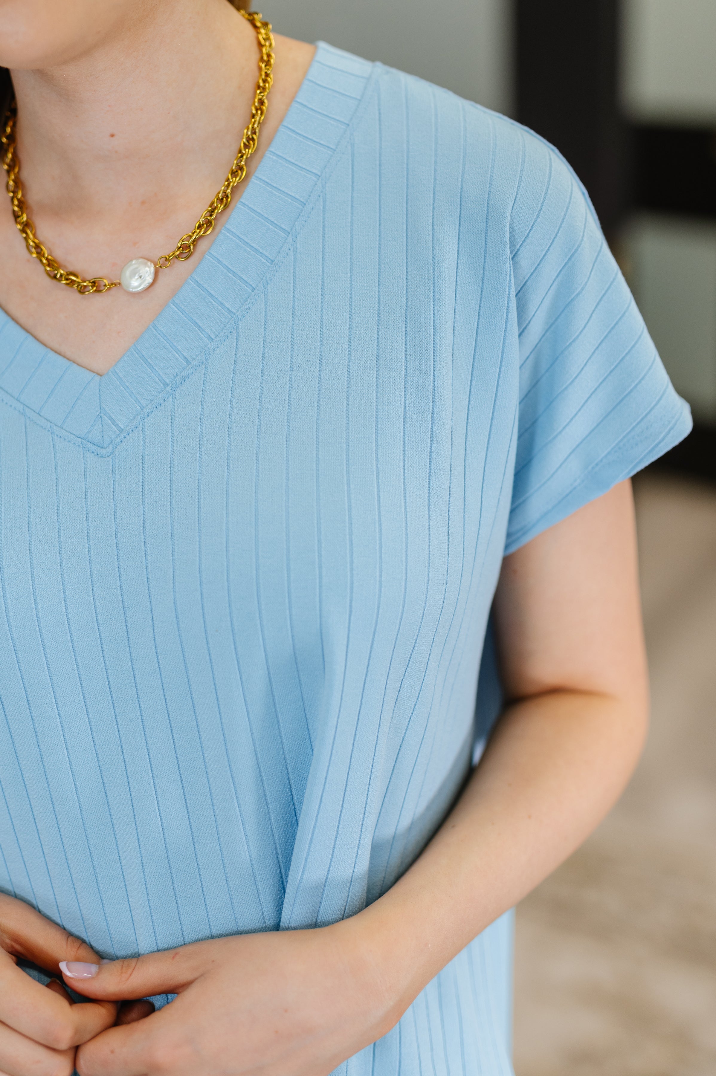 Close-up of a woman wearing a sky blue ribbed V-neck top with short sleeves and a gold chain necklace with a pearl pendant