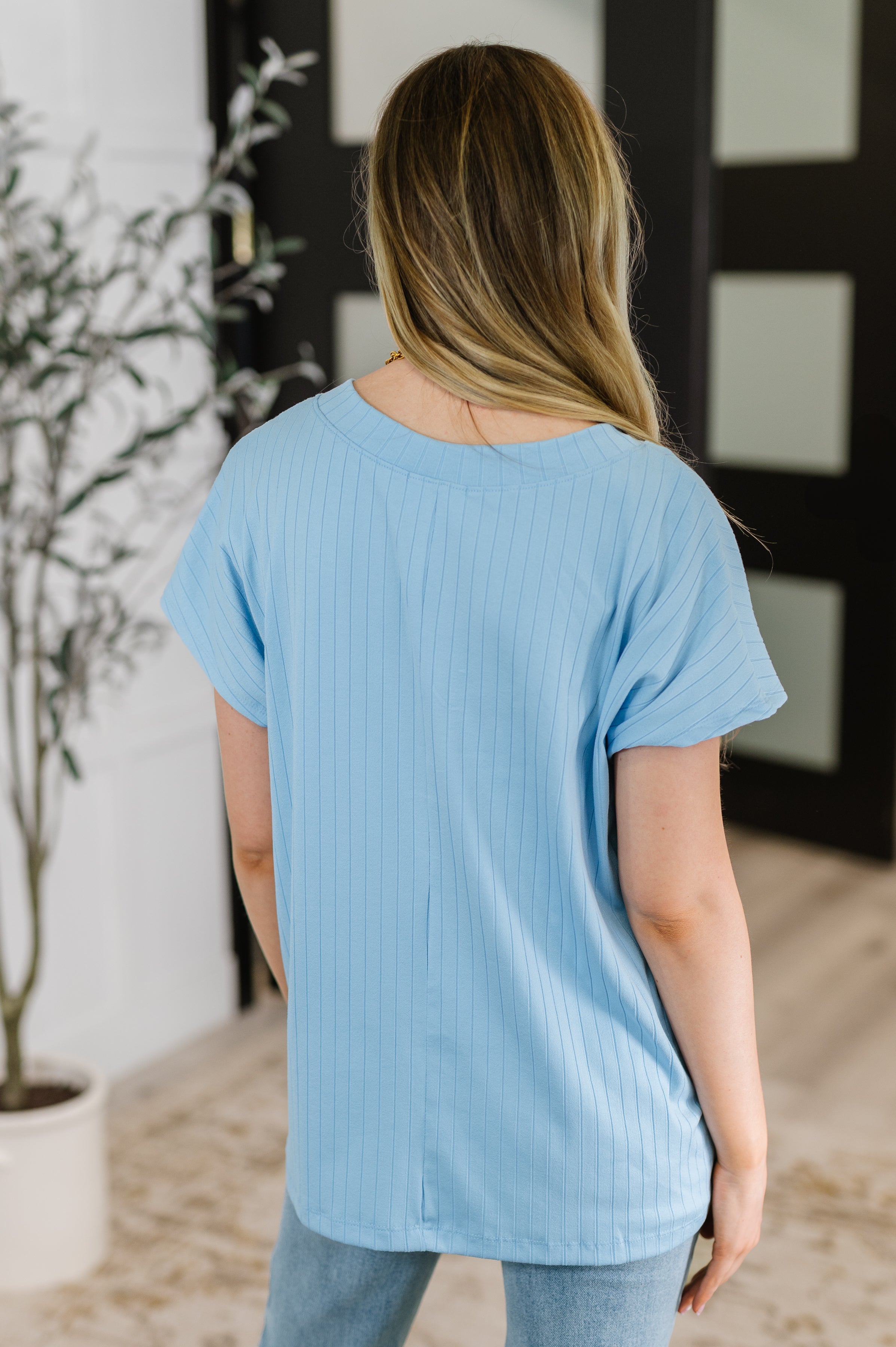 Woman wearing a sky blue ribbed short sleeve top with a relaxed fit, shown from behind in an indoor setting