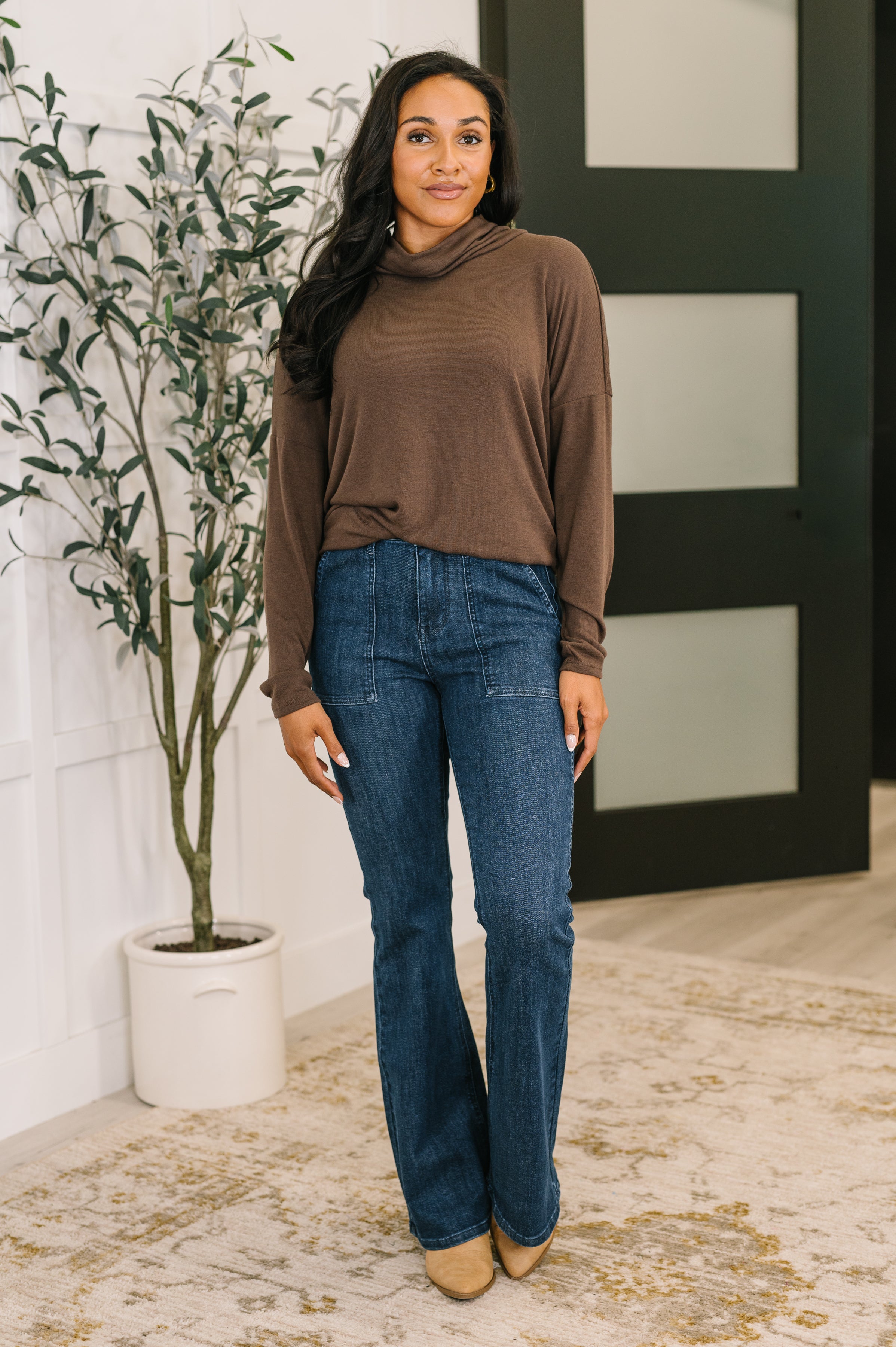 Woman wearing a chocolate cowl neck pullover paired with high-waist blue jeans and tan boots indoors by plant and door