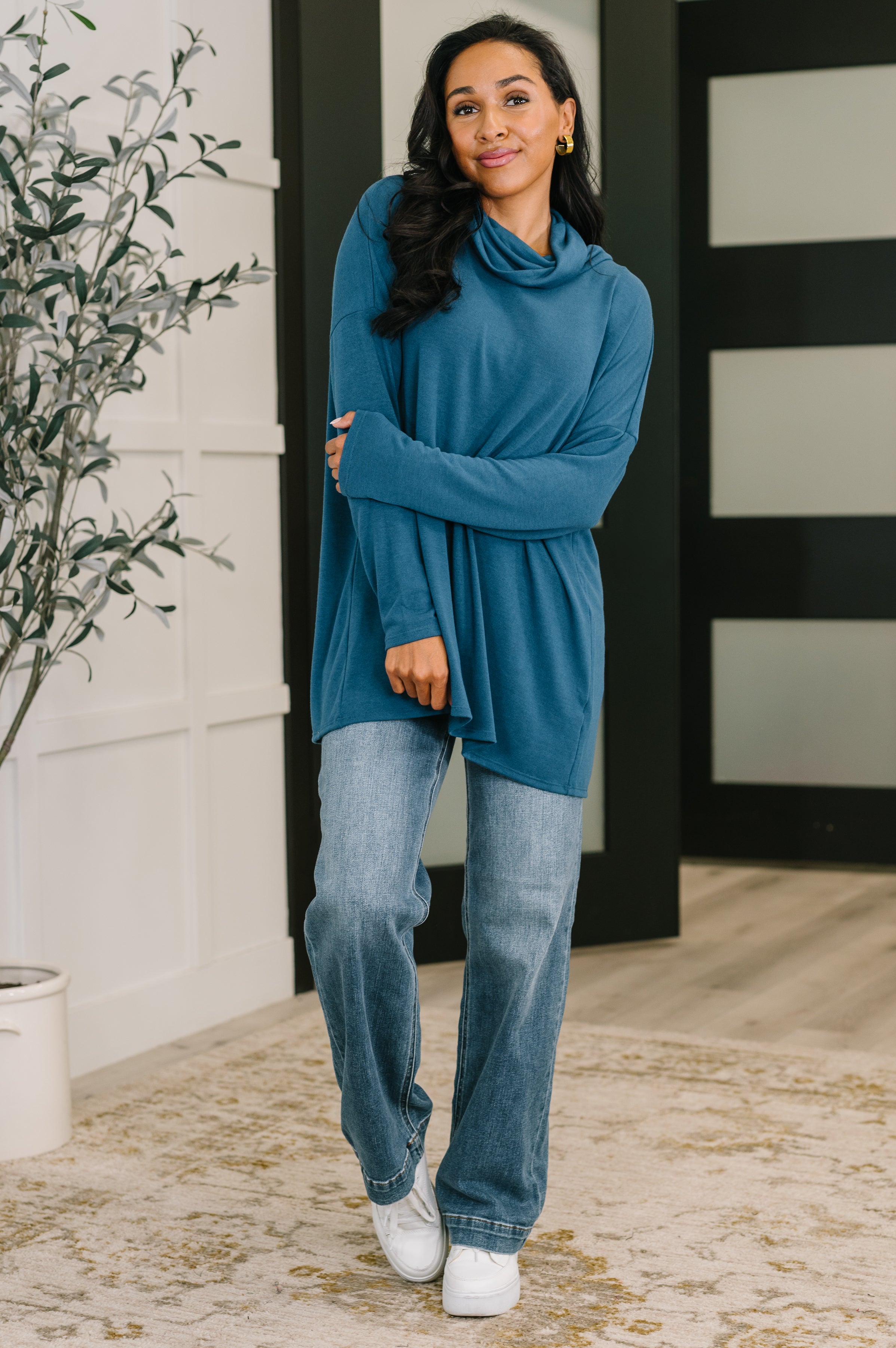 Woman wearing a dark teal cowl neck pullover with long sleeves paired with light wash jeans and white sneakers indoors