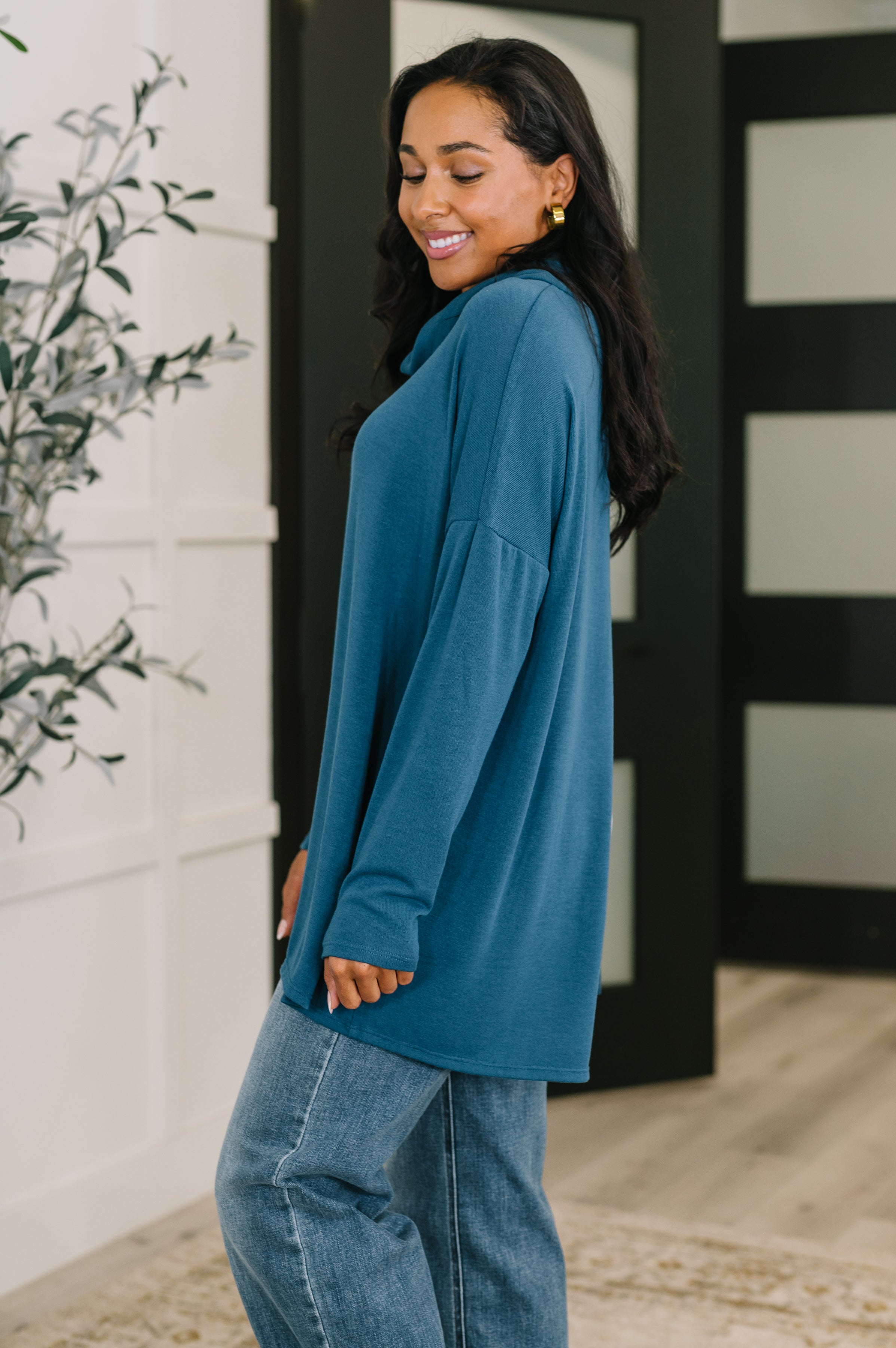 Smiling woman wearing a loose dark teal cowl neck pullover with long sleeves and casual blue jeans indoors