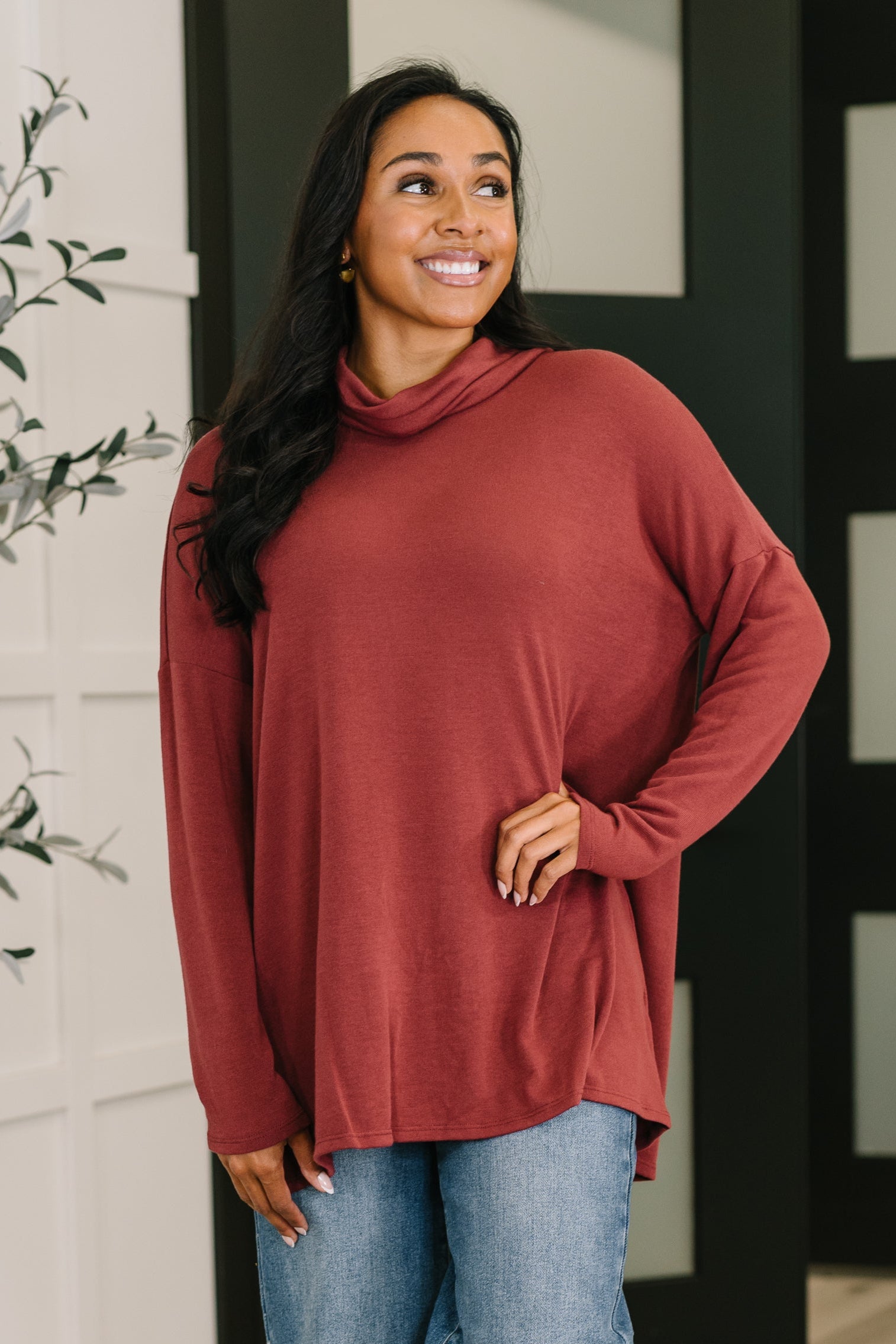 Smiling woman wearing a relaxed fit wine-colored cowl neck pullover paired with light wash jeans indoors