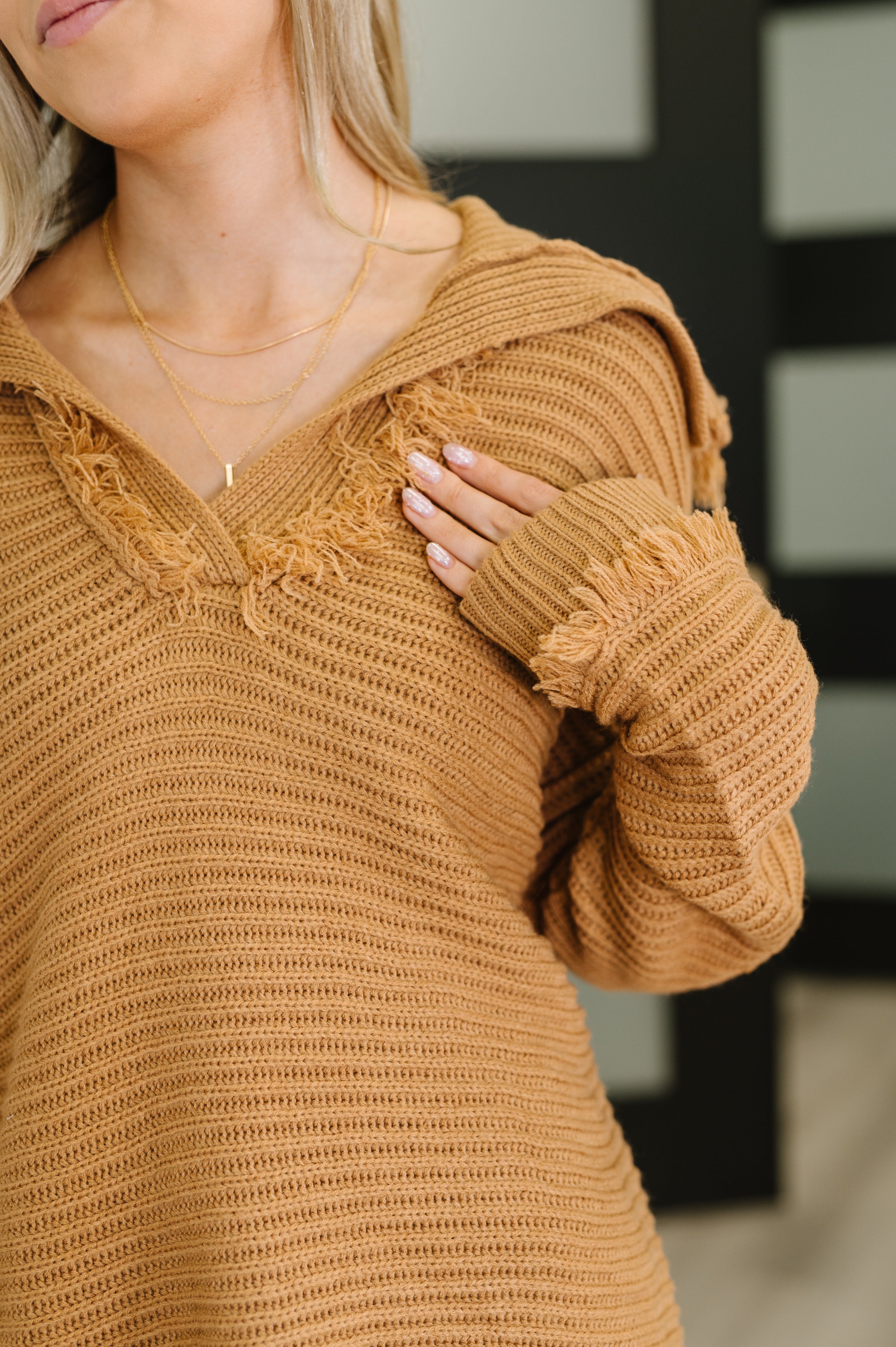 Close-up of a woman wearing a camel knit sweater with frayed V-neck and sleeve cuffs, accessorized with layered gold necklaces