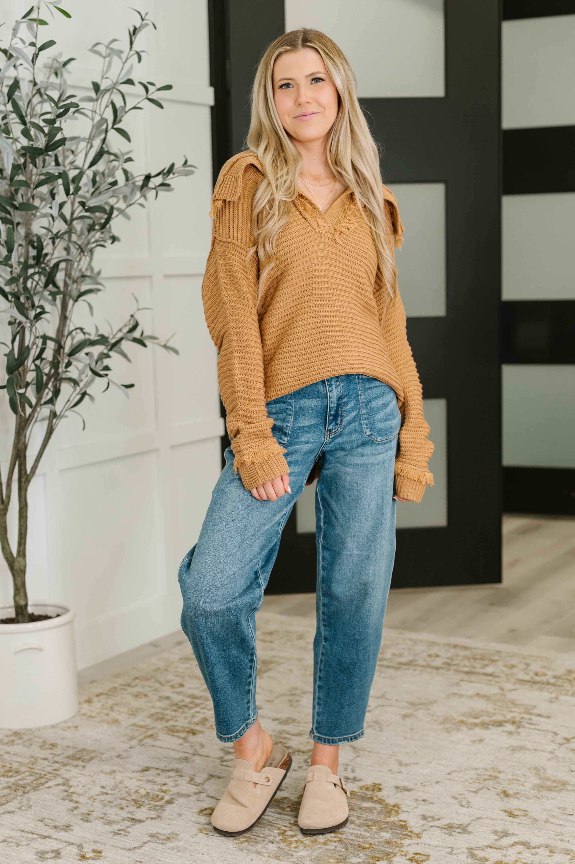 Woman wearing a camel ribbed knit sweater with button shoulder details paired with relaxed blue jeans indoors
