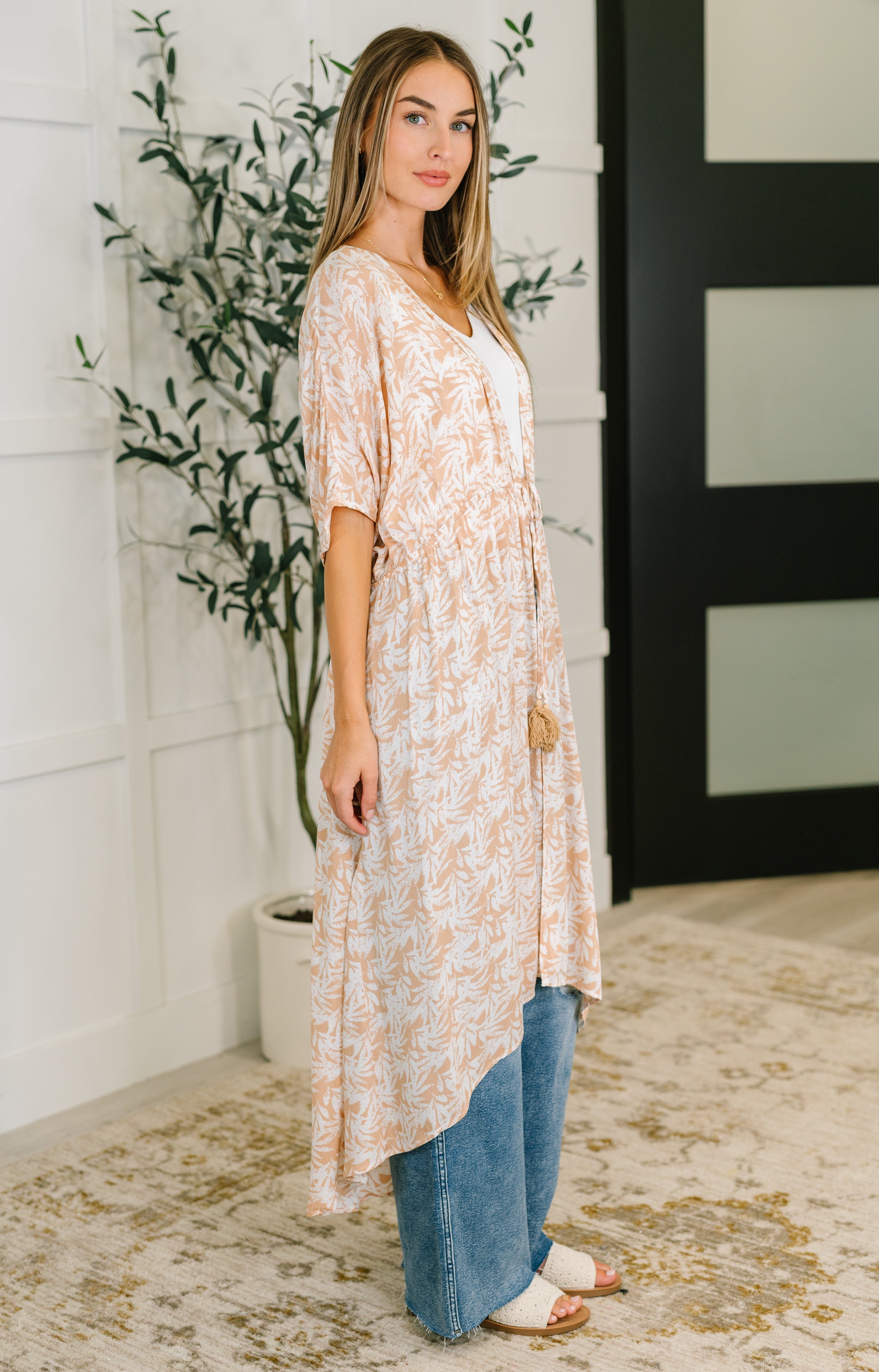 Woman wearing rust tropical leaf patterned lightweight gauze kimono with elastic waist tie over white top and blue jeans indoors