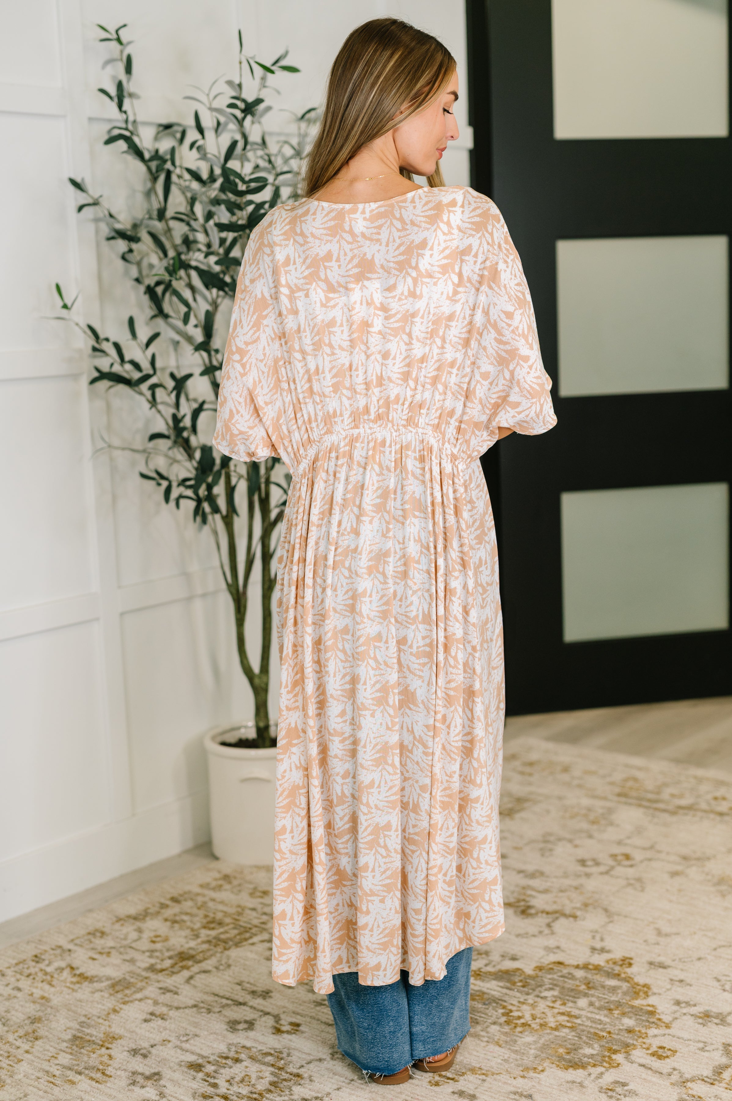 Woman wearing rust tropical leaf patterned lightweight kimono with elastic waist tie and blue jeans standing indoors facing away