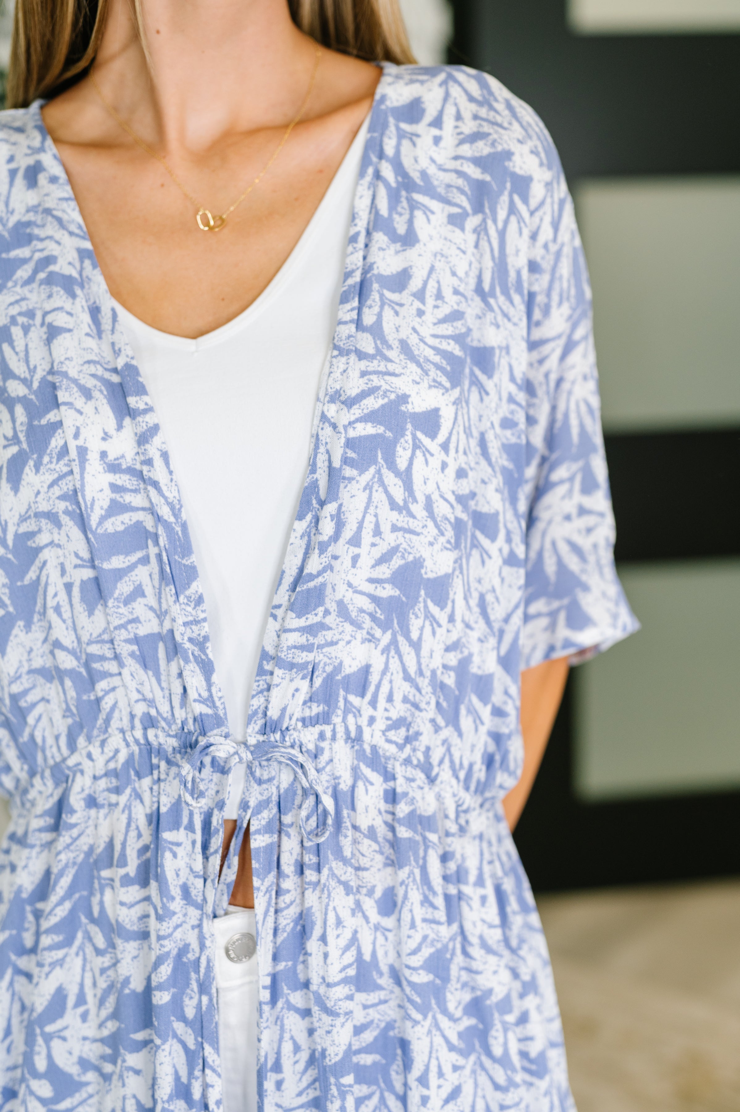 Close-up of a woman wearing a periwinkle tropical print lightweight kimono with elastic waist tie over white top and pants indoors