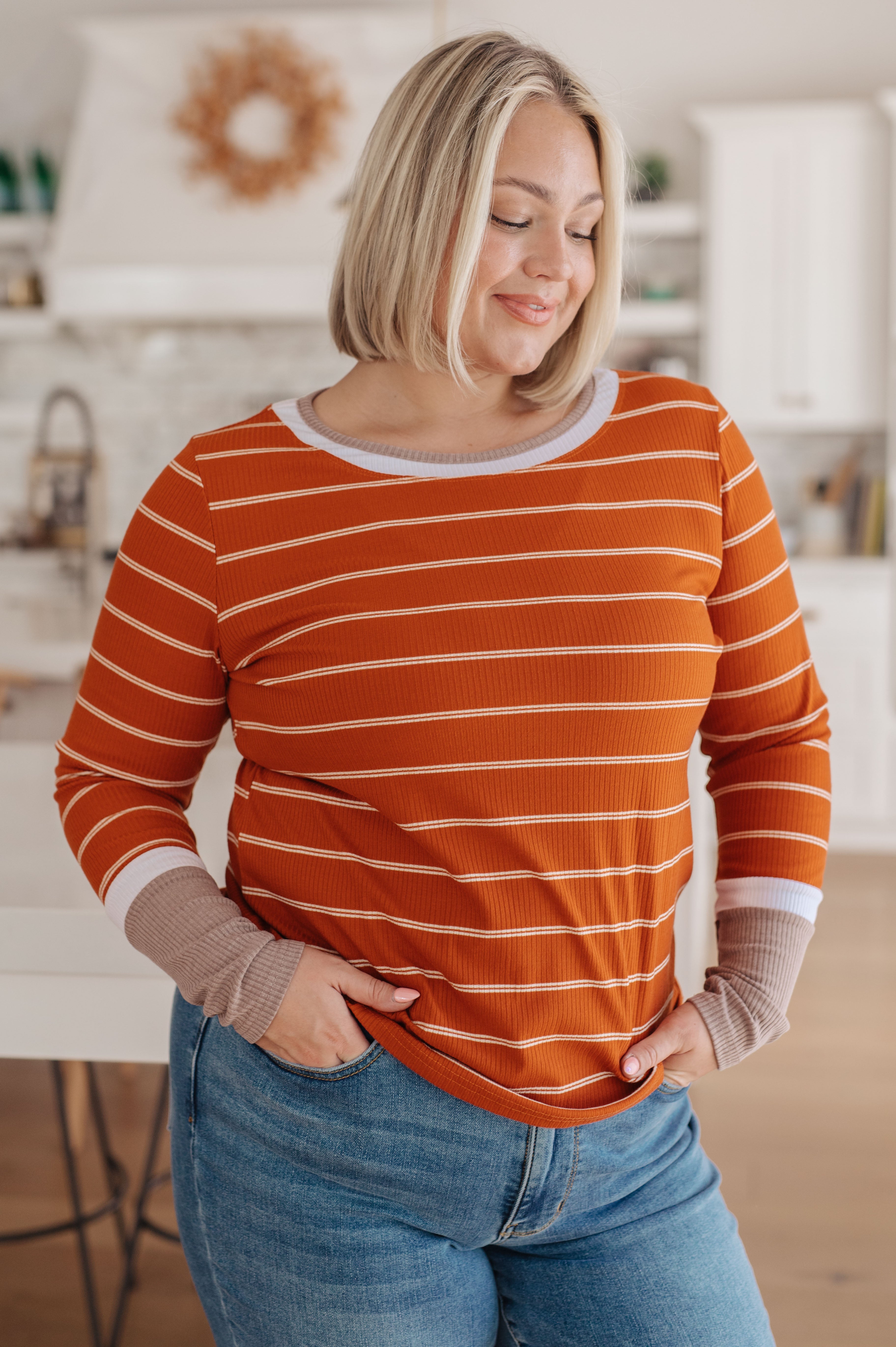Woman wearing an orange striped long sleeve tee with layered cuffs paired with blue jeans in a cozy kitchen setting