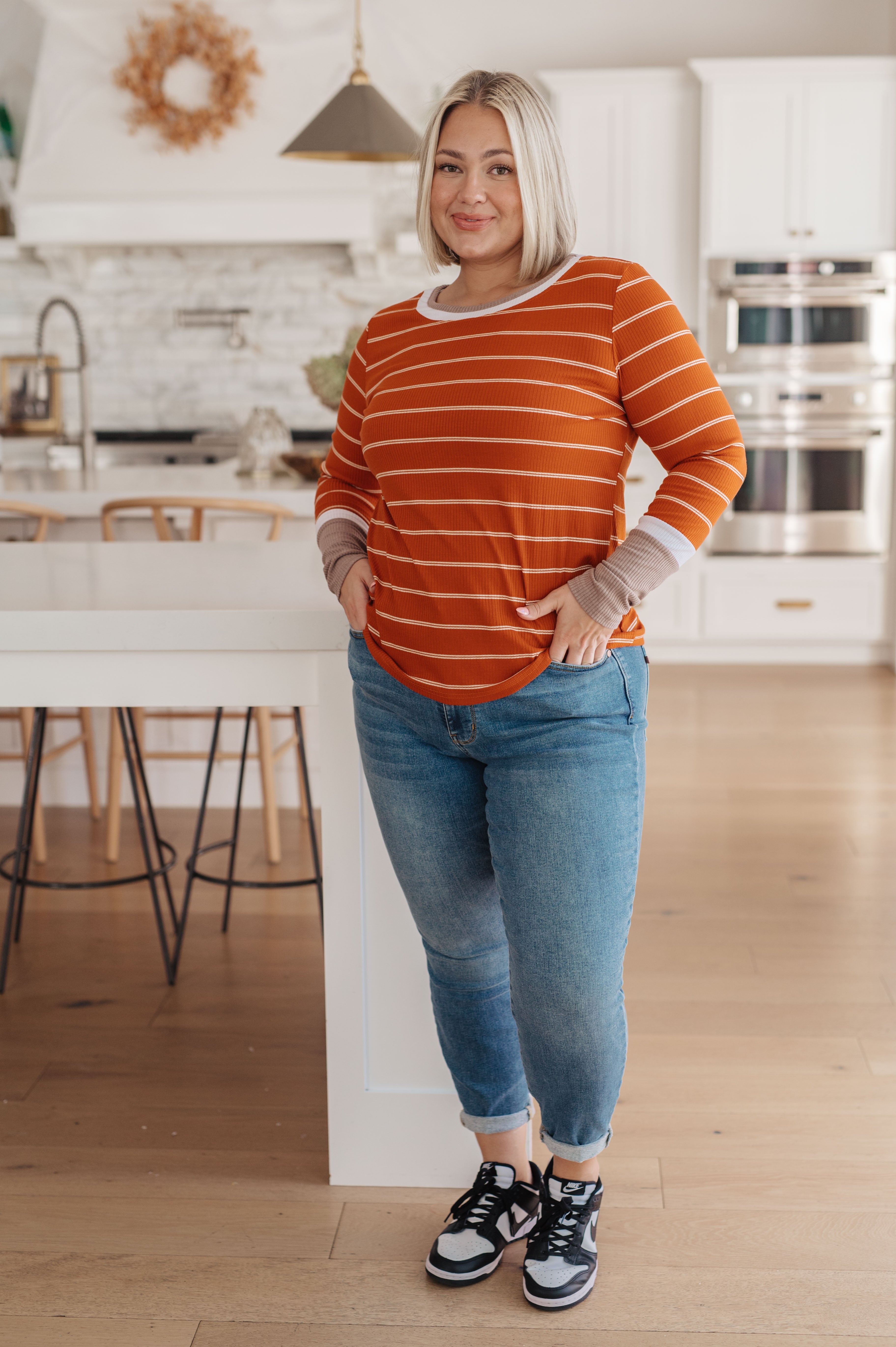 Woman wearing an orange striped long sleeve tee with grey cuffs paired with casual blue jeans and sneakers indoors