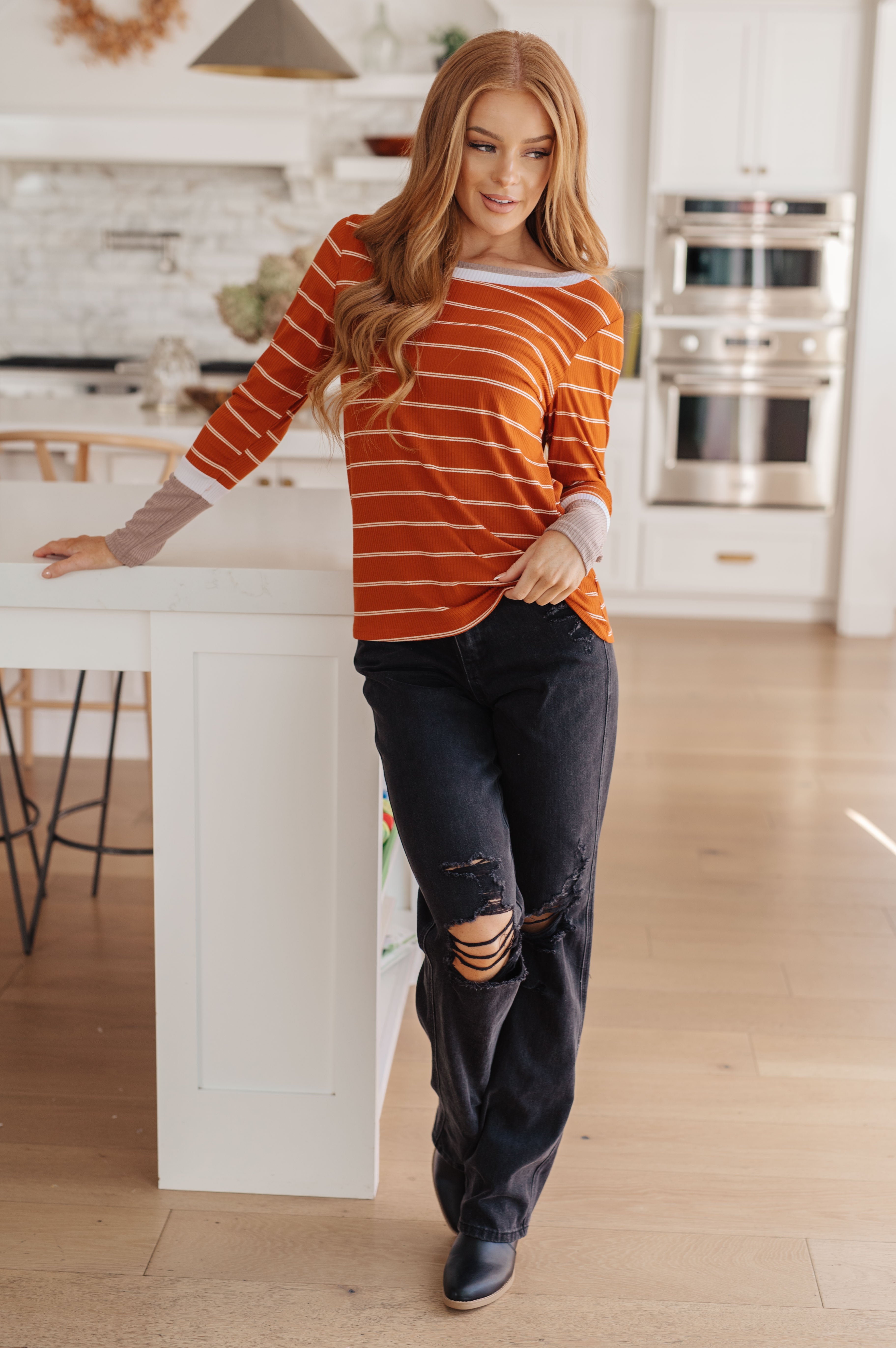 Woman in an orange striped long sleeve tee and black distressed jeans standing casually in a bright kitchen setting