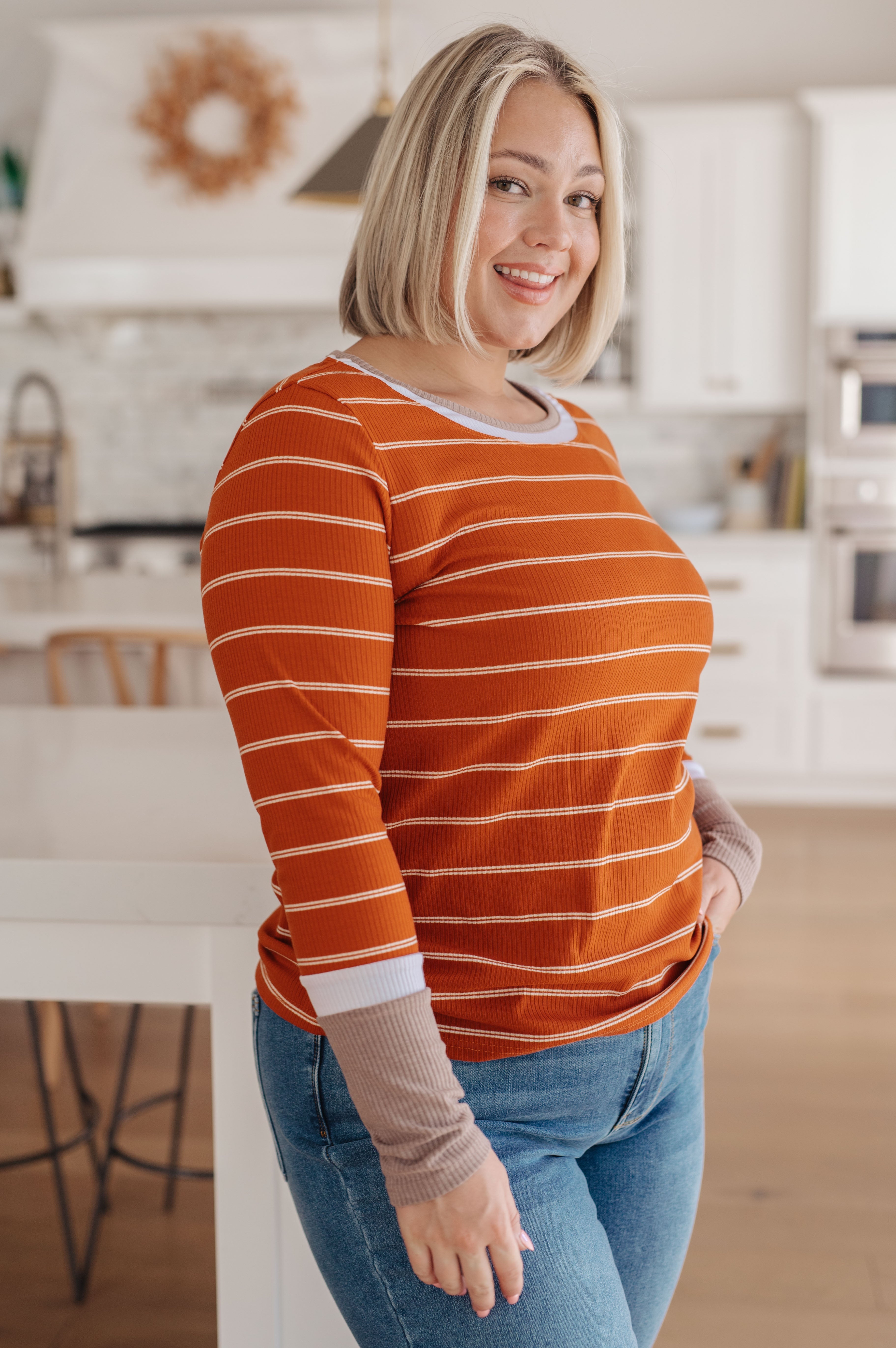 Smiling woman wearing an orange striped long sleeve tee with contrasting cuffs and casual blue jeans in a cozy kitchen