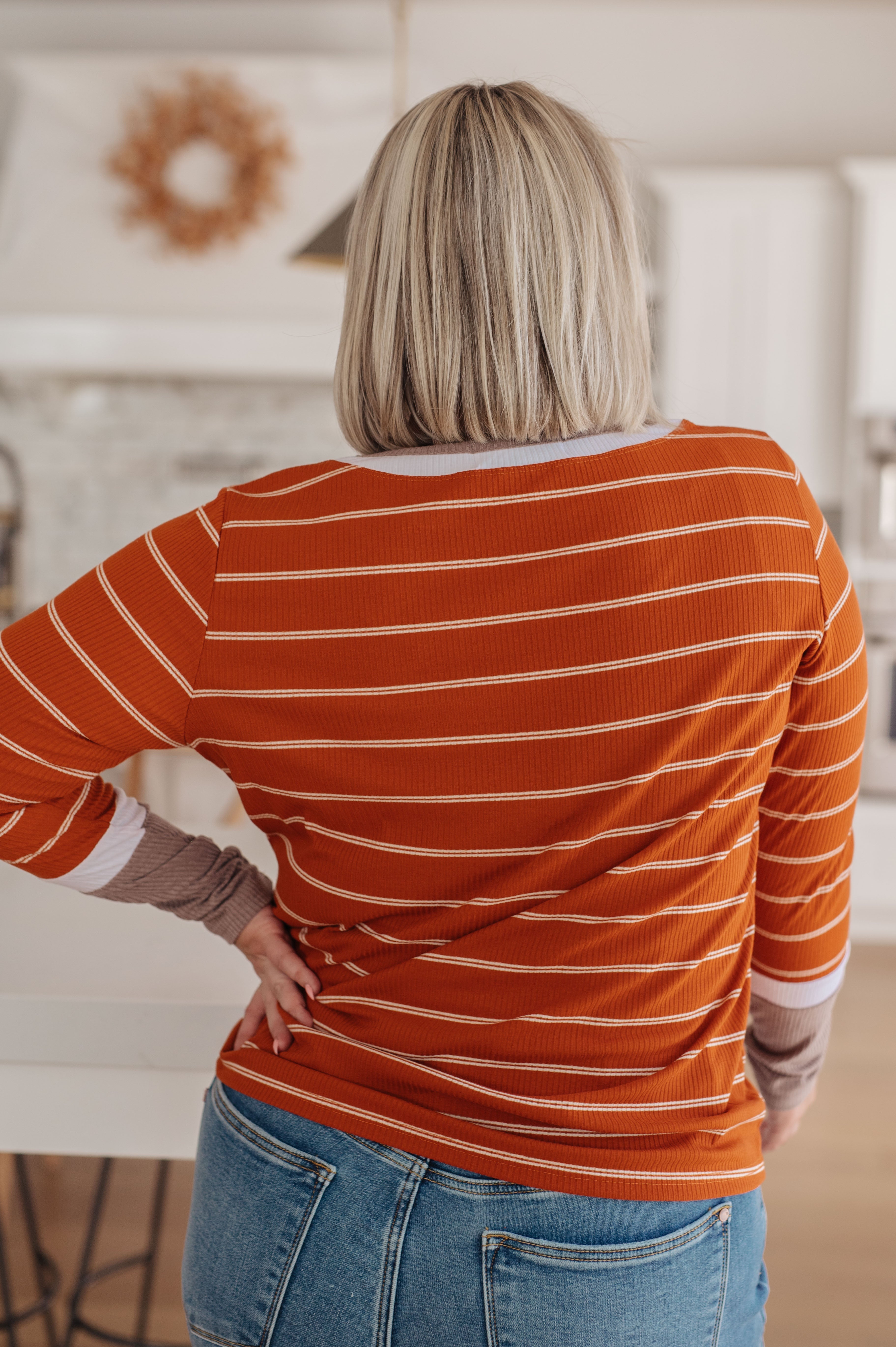 Woman seen from behind wearing an orange striped long sleeve tee with layered cuffs and fitted blue jeans indoors