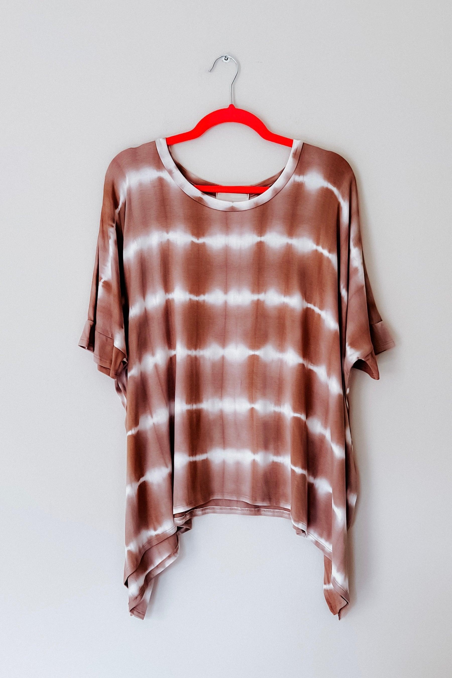 Loose-fitting striped tie dye tunic in muted earth tones hanging on a bright red hanger against a plain wall